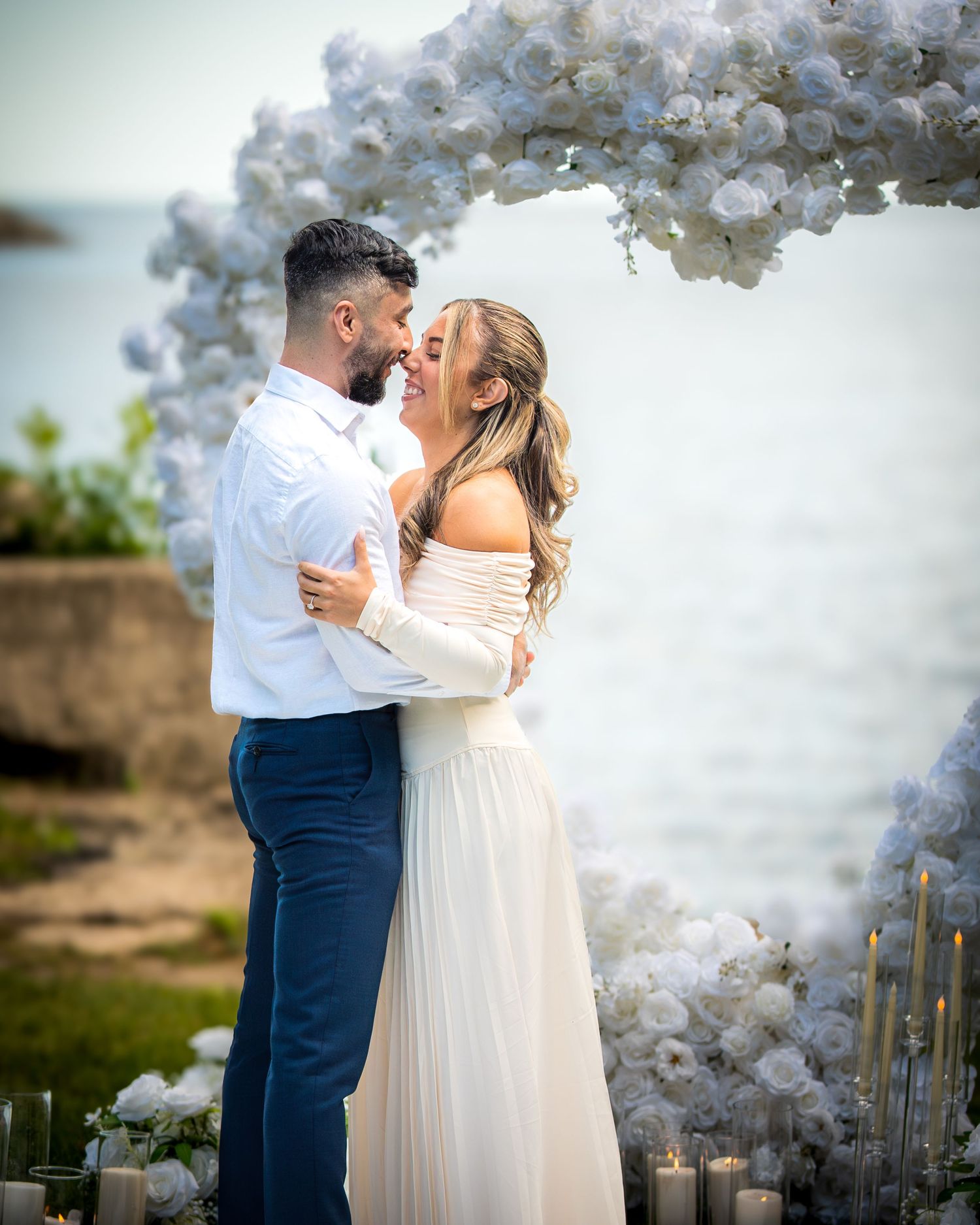 A couple shares an intimate moment under an elegant white balloon arch overlooking the sea.