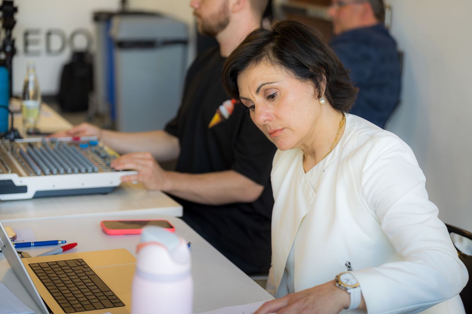 Woman in white blazer working on laptop at office desk with colleague in background.