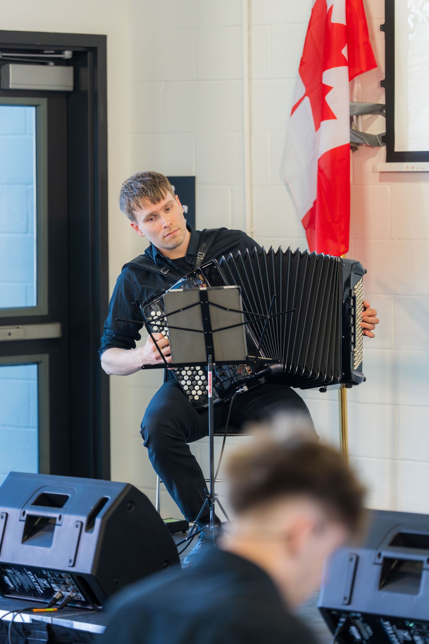 Male musician playing black accordion on stage with Canadian flag in background.