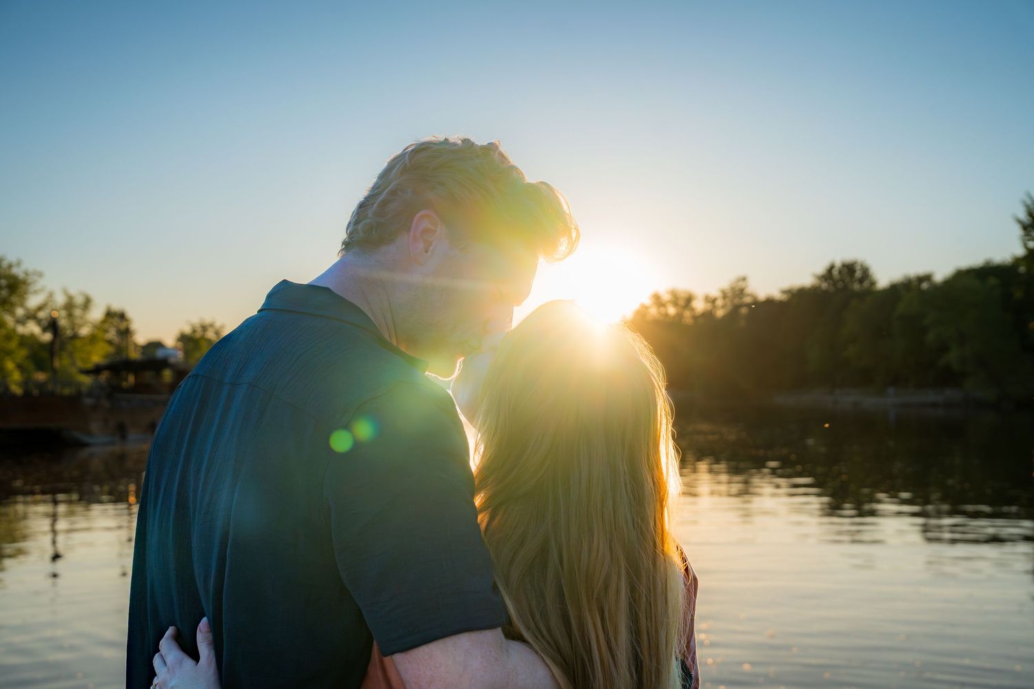 Romantic couple embracing by a lake at sunset with golden lens flare.
