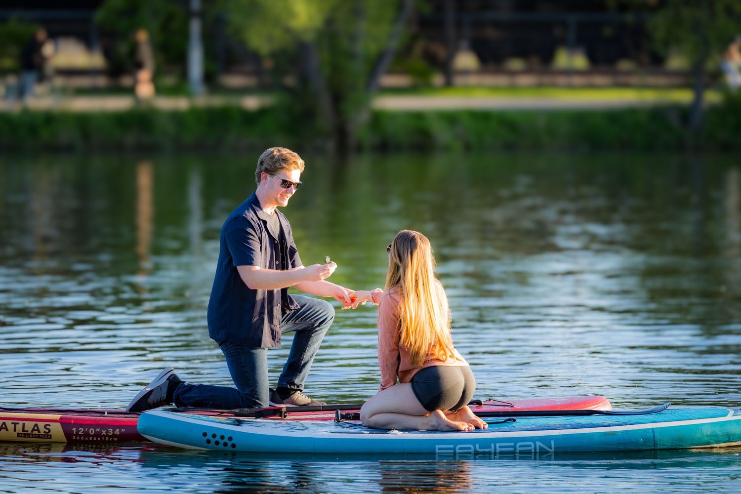 Man proposing to woman on a paddleboard on a calm lake surrounded by trees.