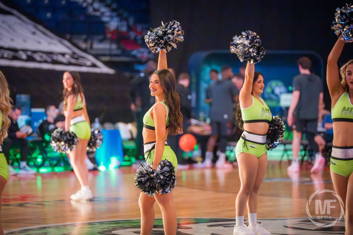 Cheerleader in neon yellow uniform smiles while performing with black pom-poms on a basketball court.