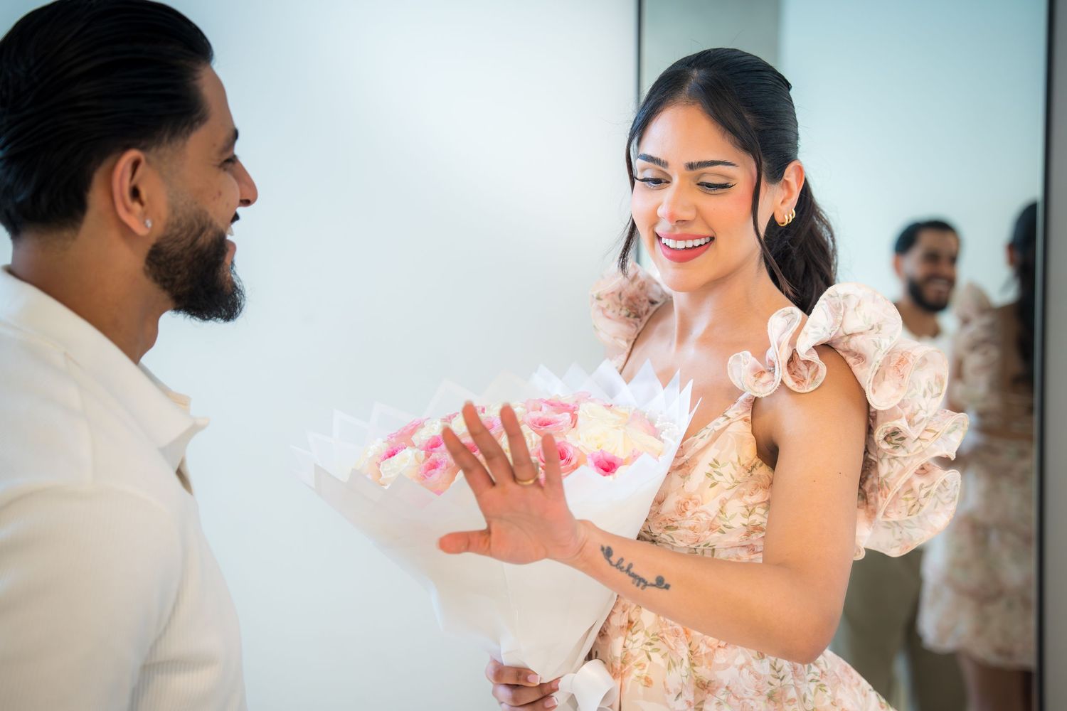 Woman in floral dress smiling while receiving pink flower bouquet from man in white outfit indoors.