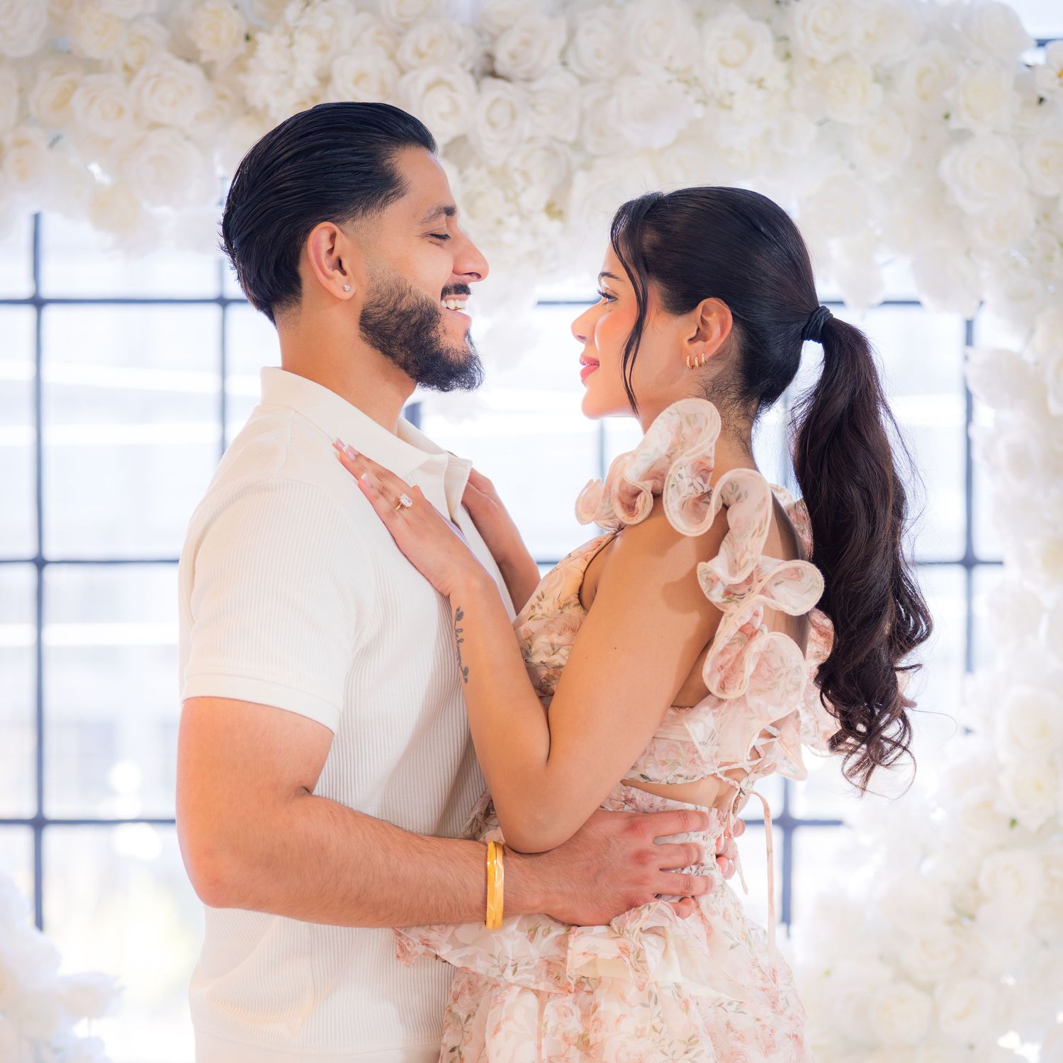 Smiling couple gazing at each other surrounded by a white floral heart arch.