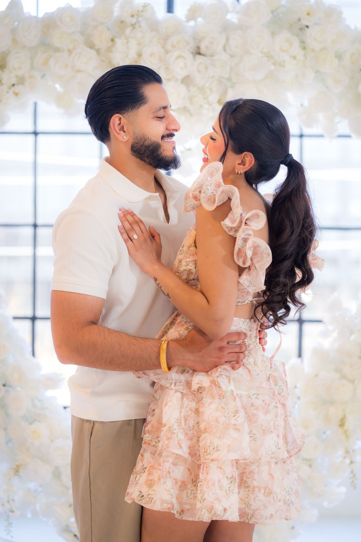 Happy couple embracing and smiling at each other in front of a white floral arch during an engagement session.