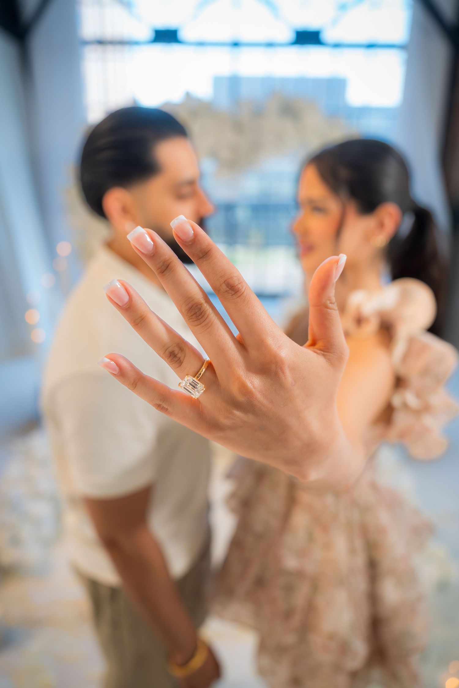 Engaged couple showing off diamond ring to camera with floral heart arch in background.