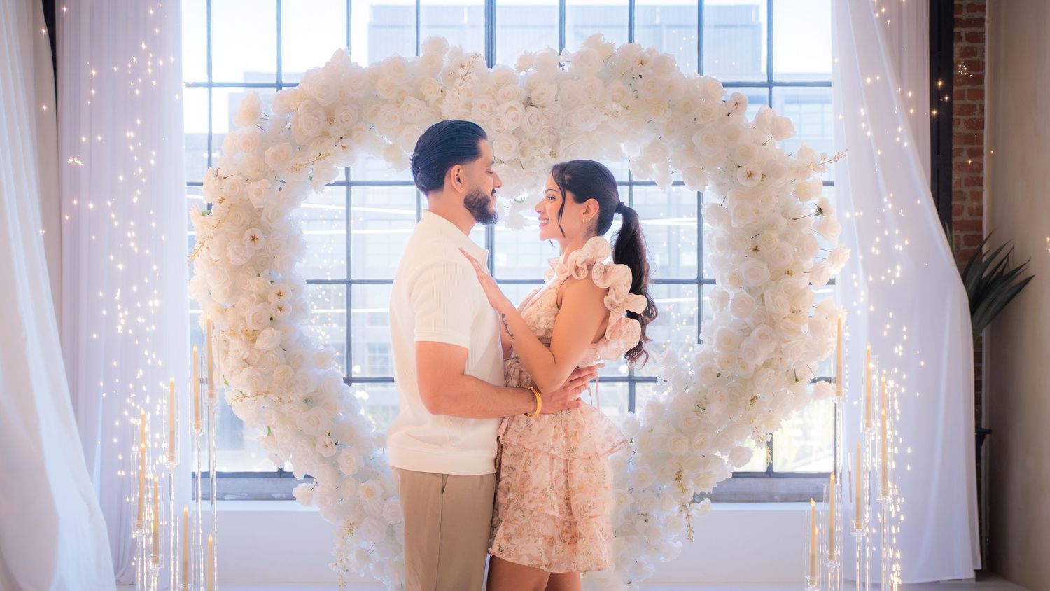 Engaged couple embracing beneath white floral heart arch with twinkling fairy lights.
