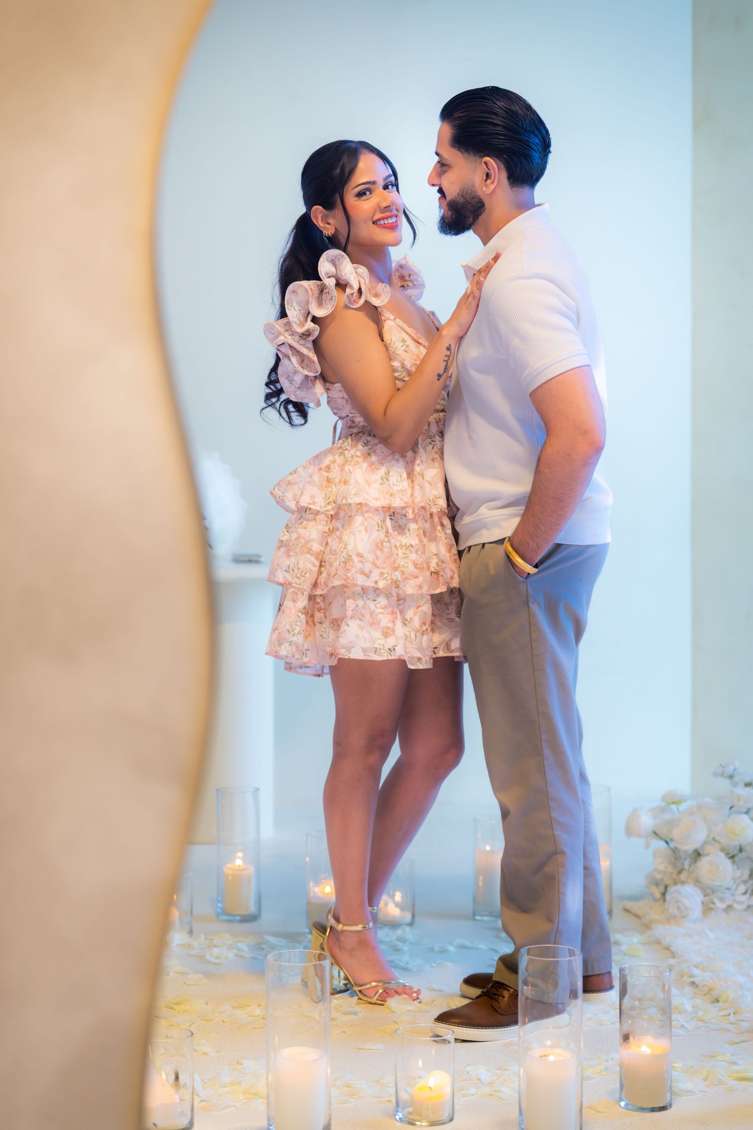 Couple standing together surrounded by candles and white floral decor in a romantic engagement setting.