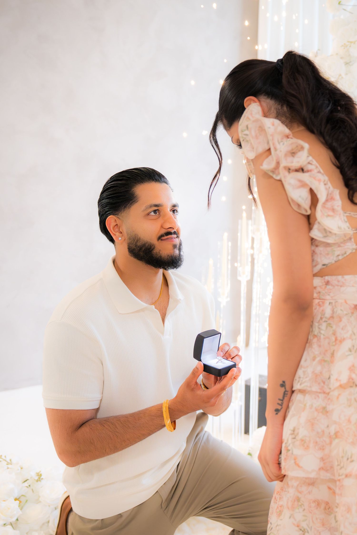 Man on one knee proposing with ring box to woman in floral dress amid romantic decor.