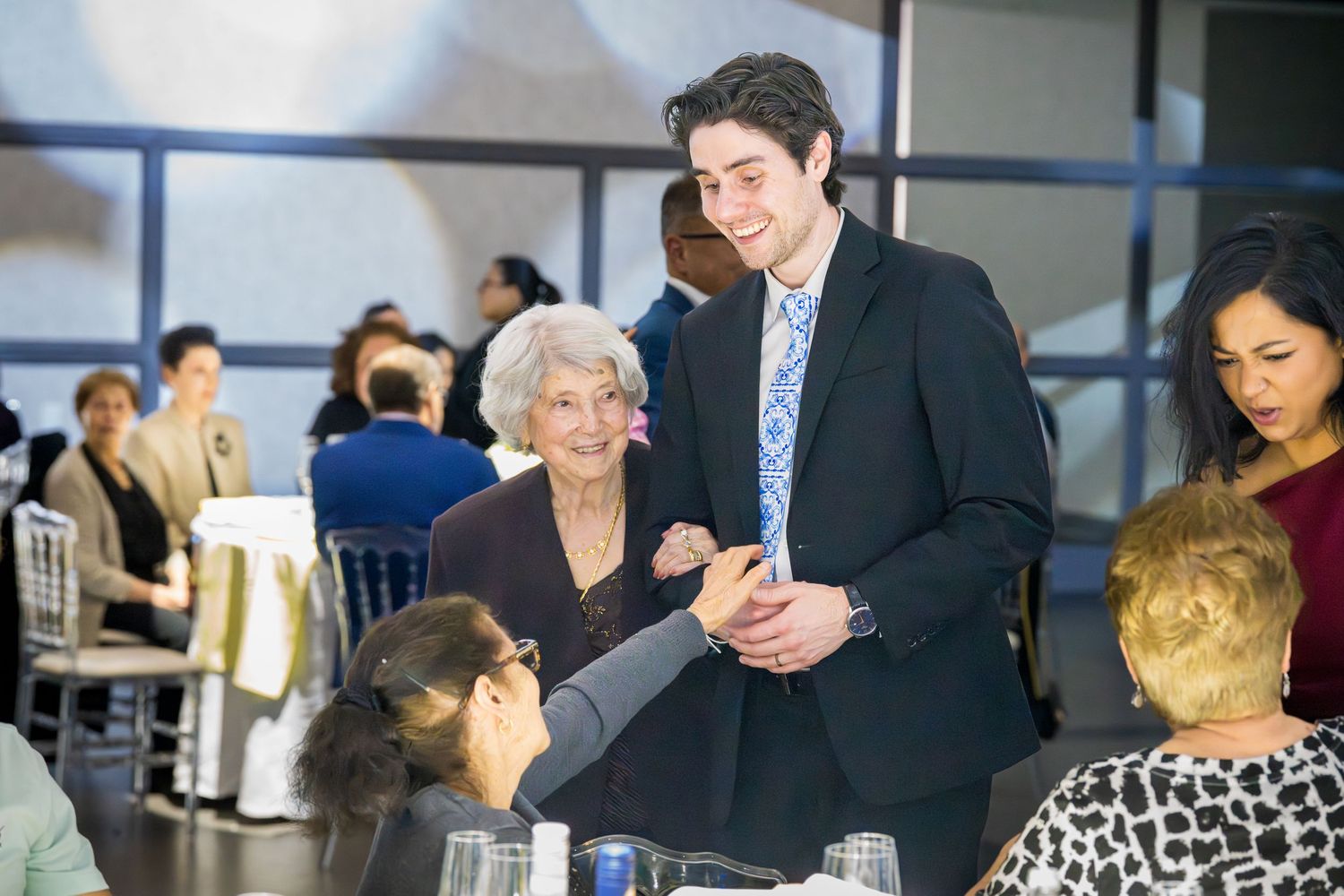 Young man in black suit greeting elderly woman at a formal event reception.