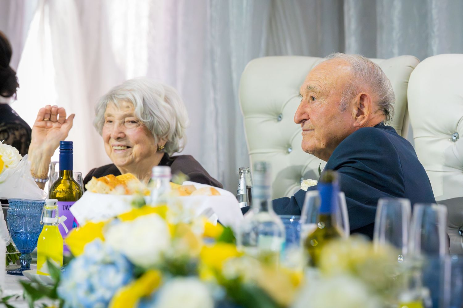 Elderly couple smiling and conversing at a festive banquet table with flowers and wine.