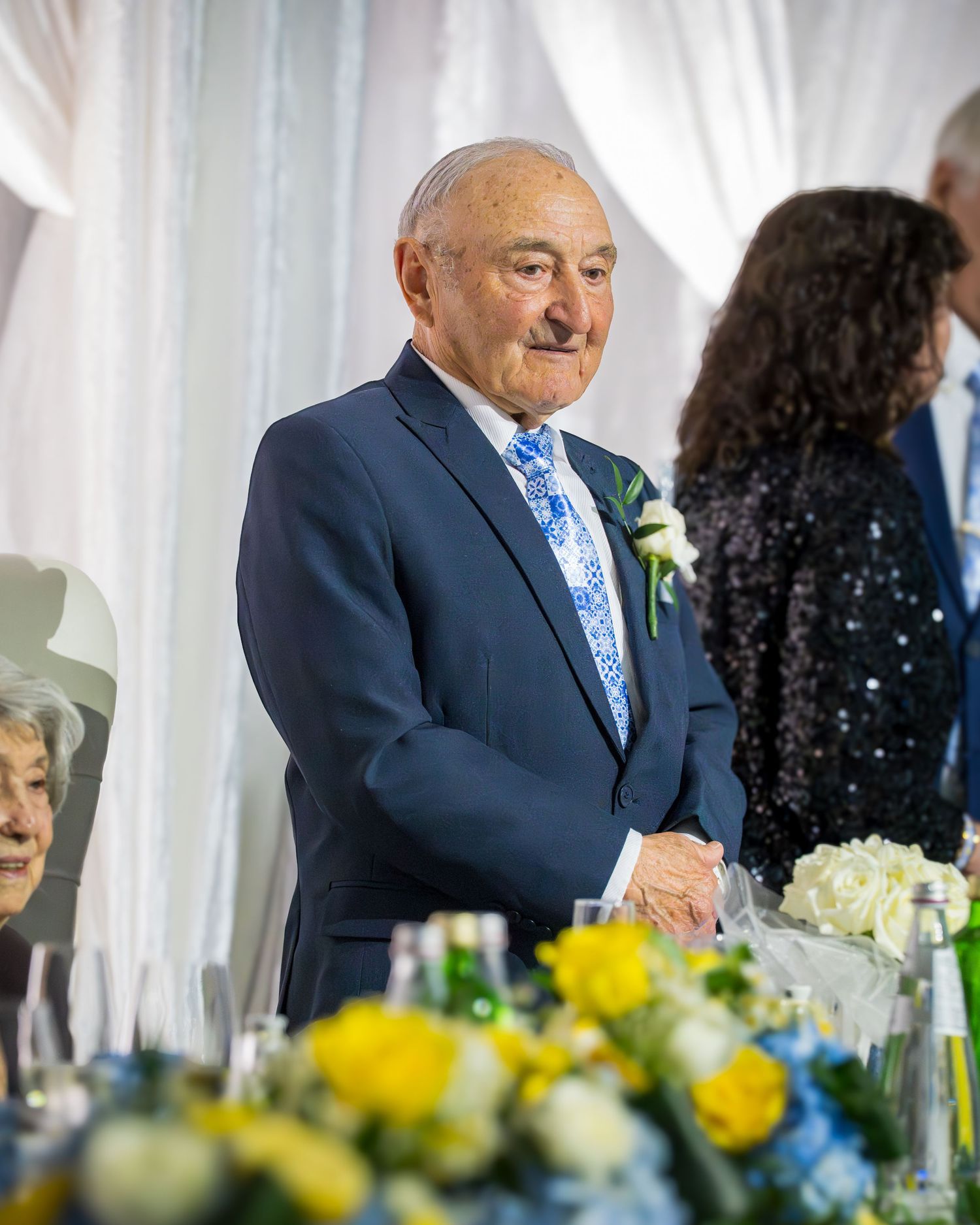 Elderly man in navy suit with floral tie and boutonniere smiling at a formal wedding reception.
