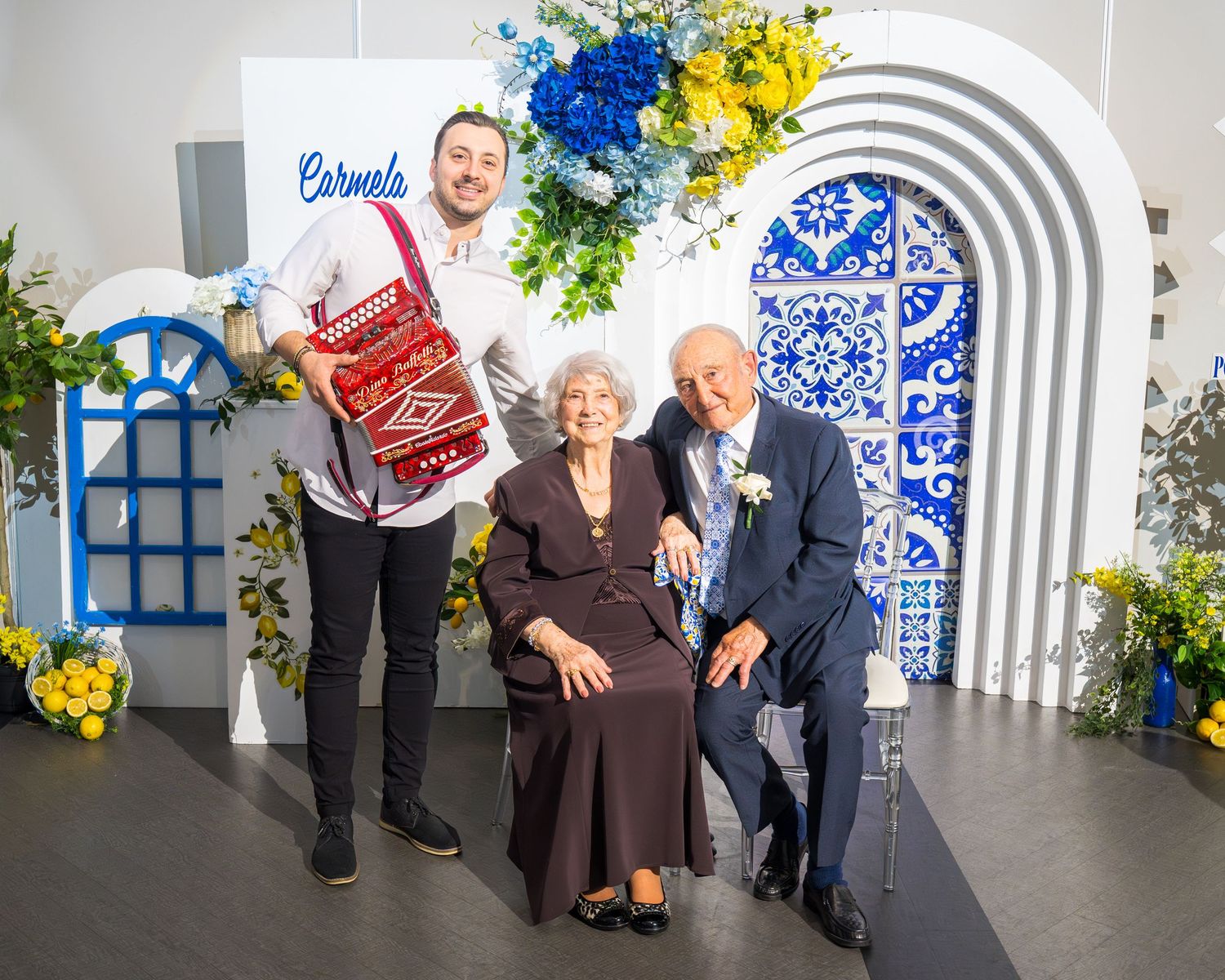 Three people posing at a decorated event backdrop, one holding an accordion.