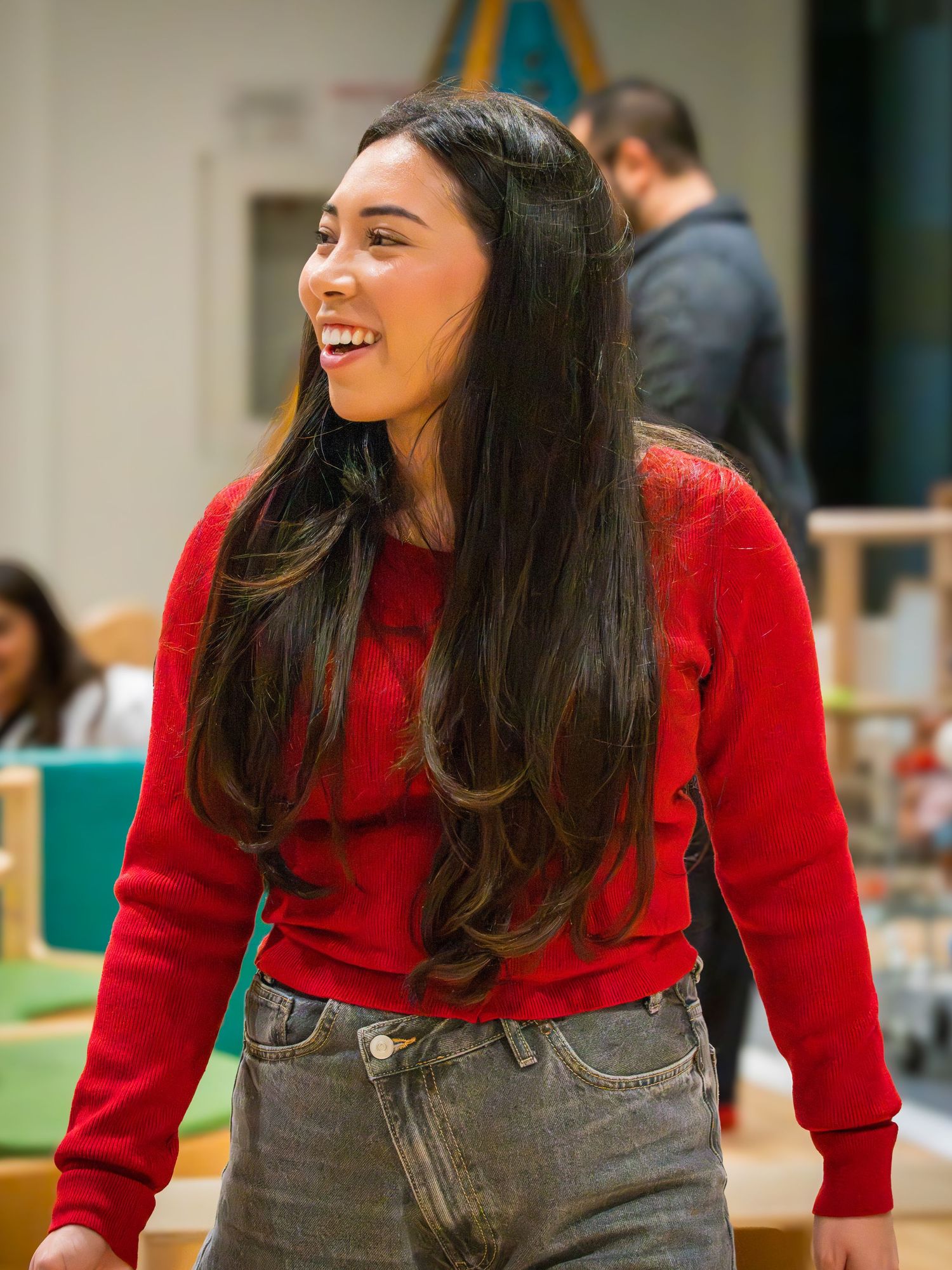 Happy young woman with long dark hair wearing a red sweater smiling in a bright indoor play space.