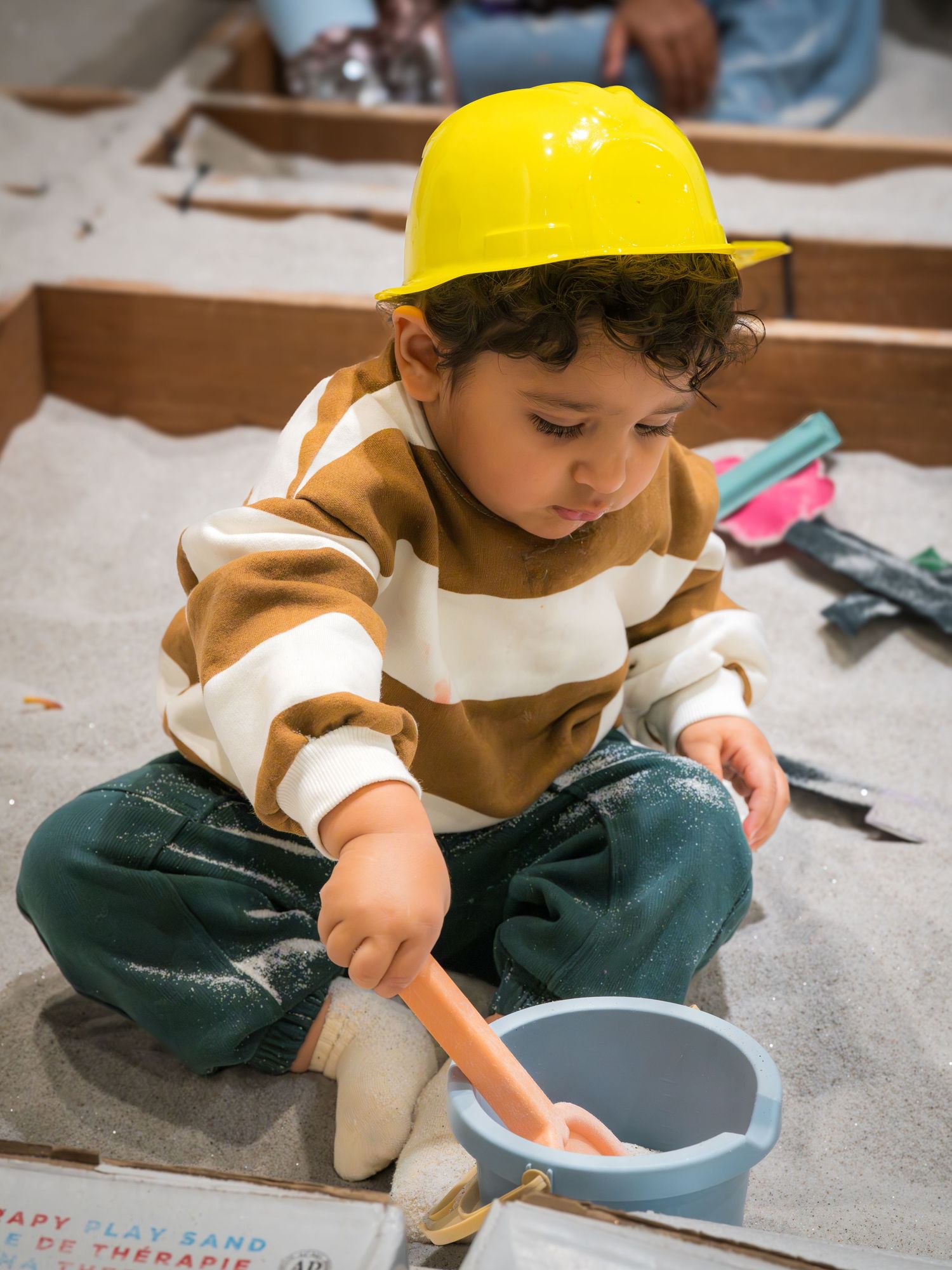 A toddler wearing a yellow hard hat sits in a sandbox scooping sand with a small bucket.