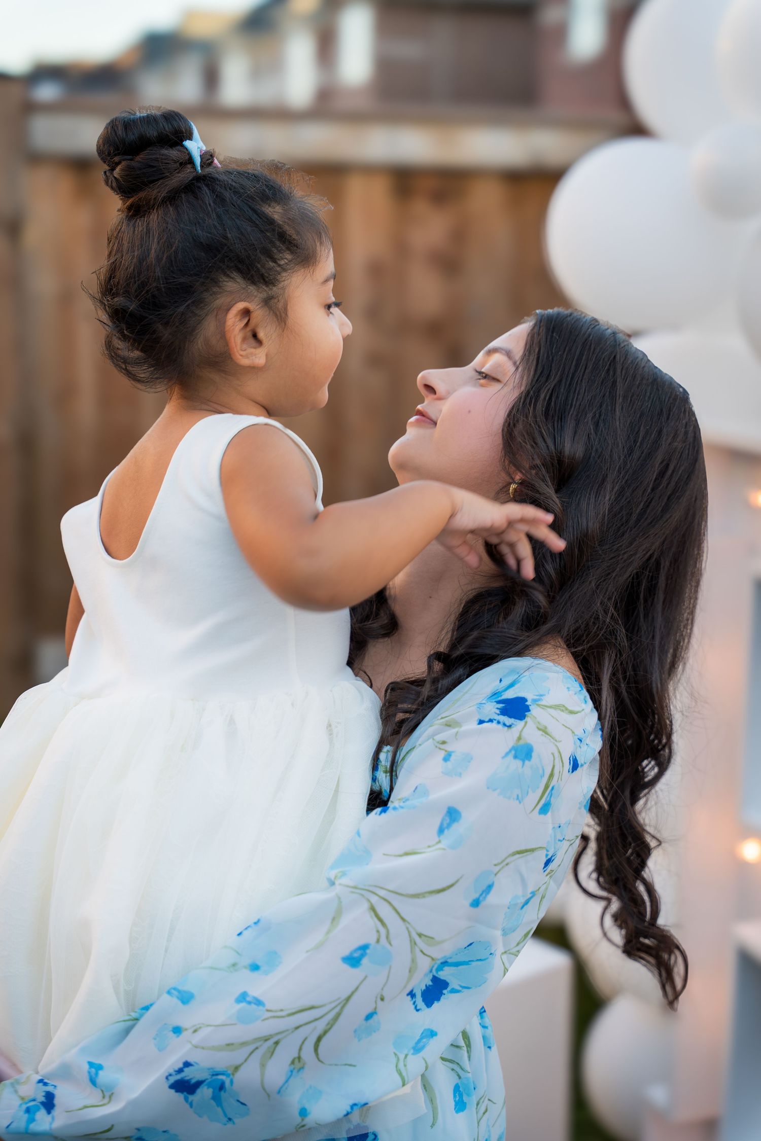 A mother tenderly holds her young daughter in a white dress at an outdoor gender reveal party.