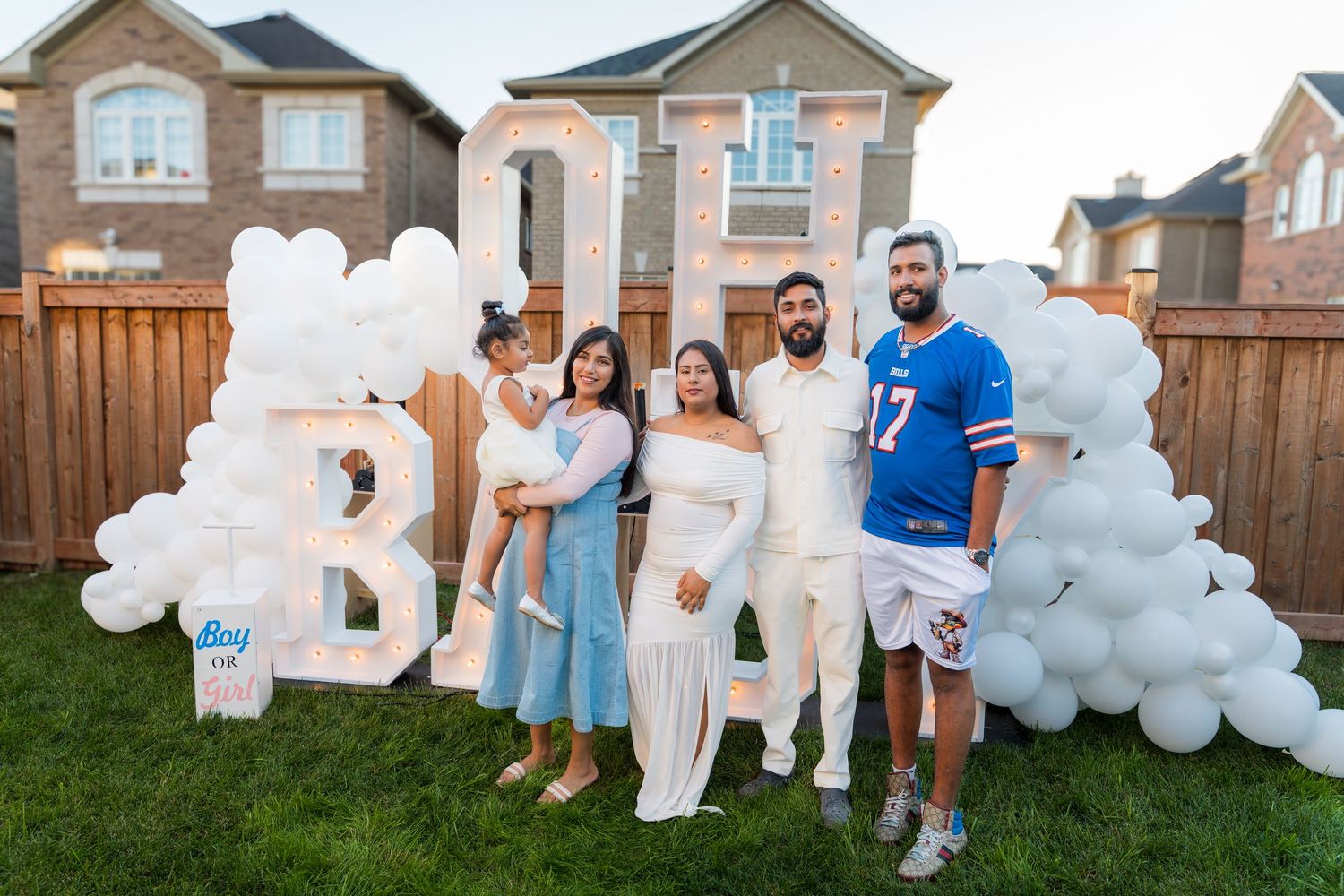 Family poses at a baby gender reveal party with large lit BABY letters and white balloon arch in backyard.