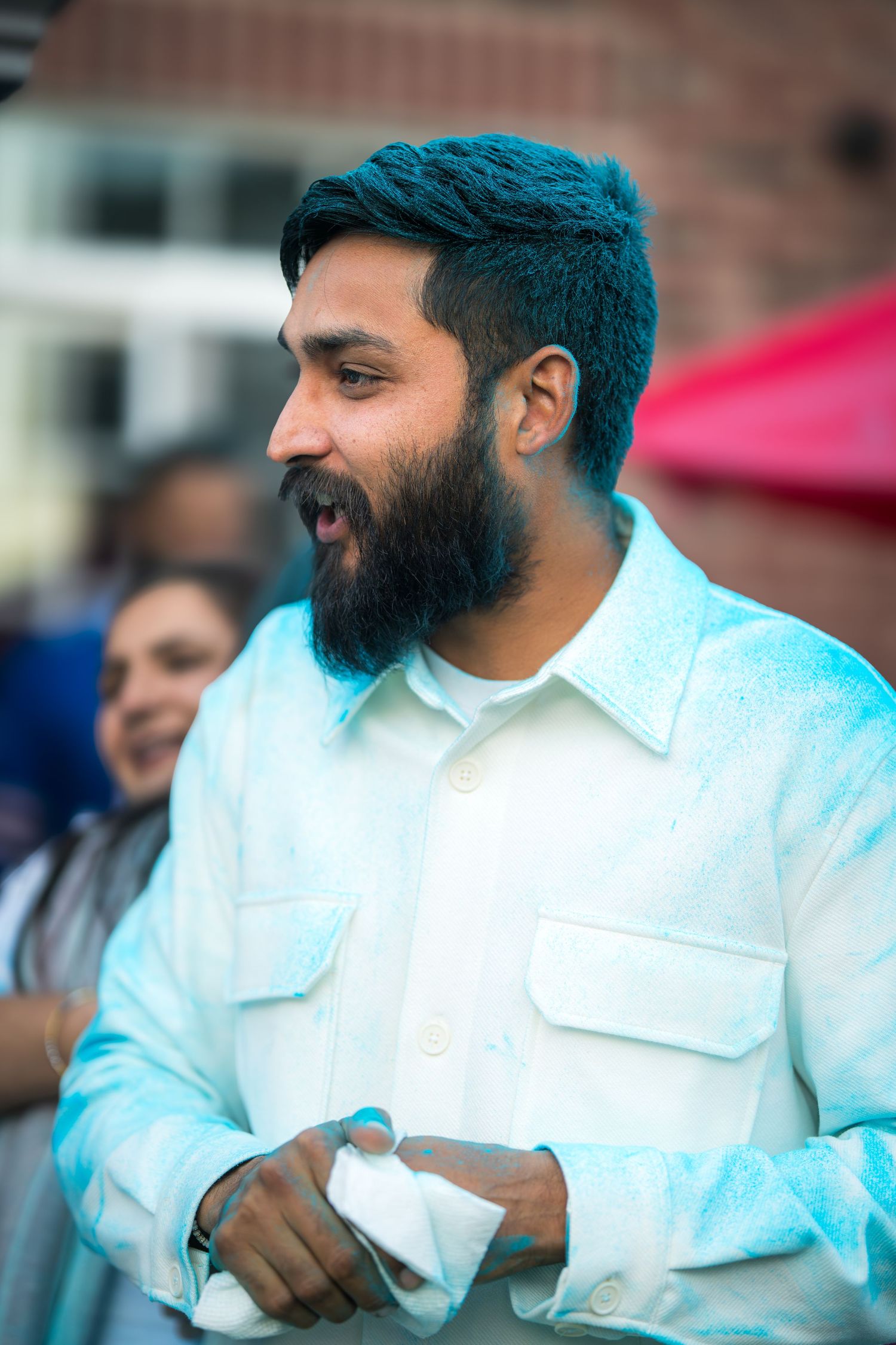 A bearded man in a white shirt covered in blue powder smiles at an outdoor Holi celebration.