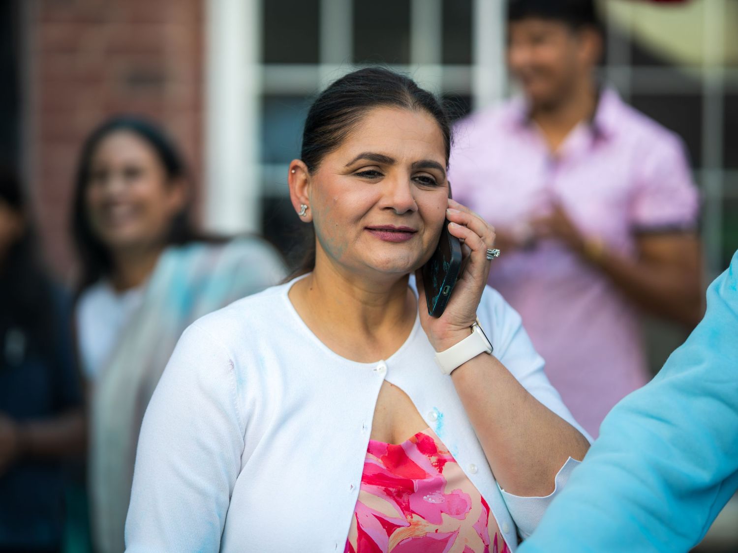 A smiling woman in a white cardigan and floral dress talking on a phone at an outdoor event.