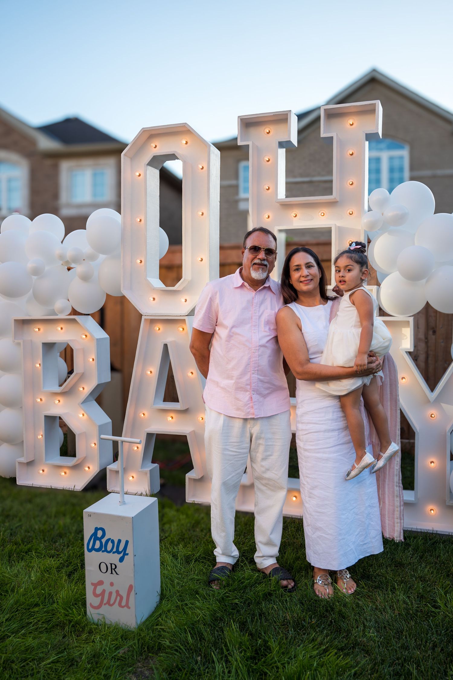 Grandparents holding child pose before lit OH BABY marquee letters with white balloon arch at night.