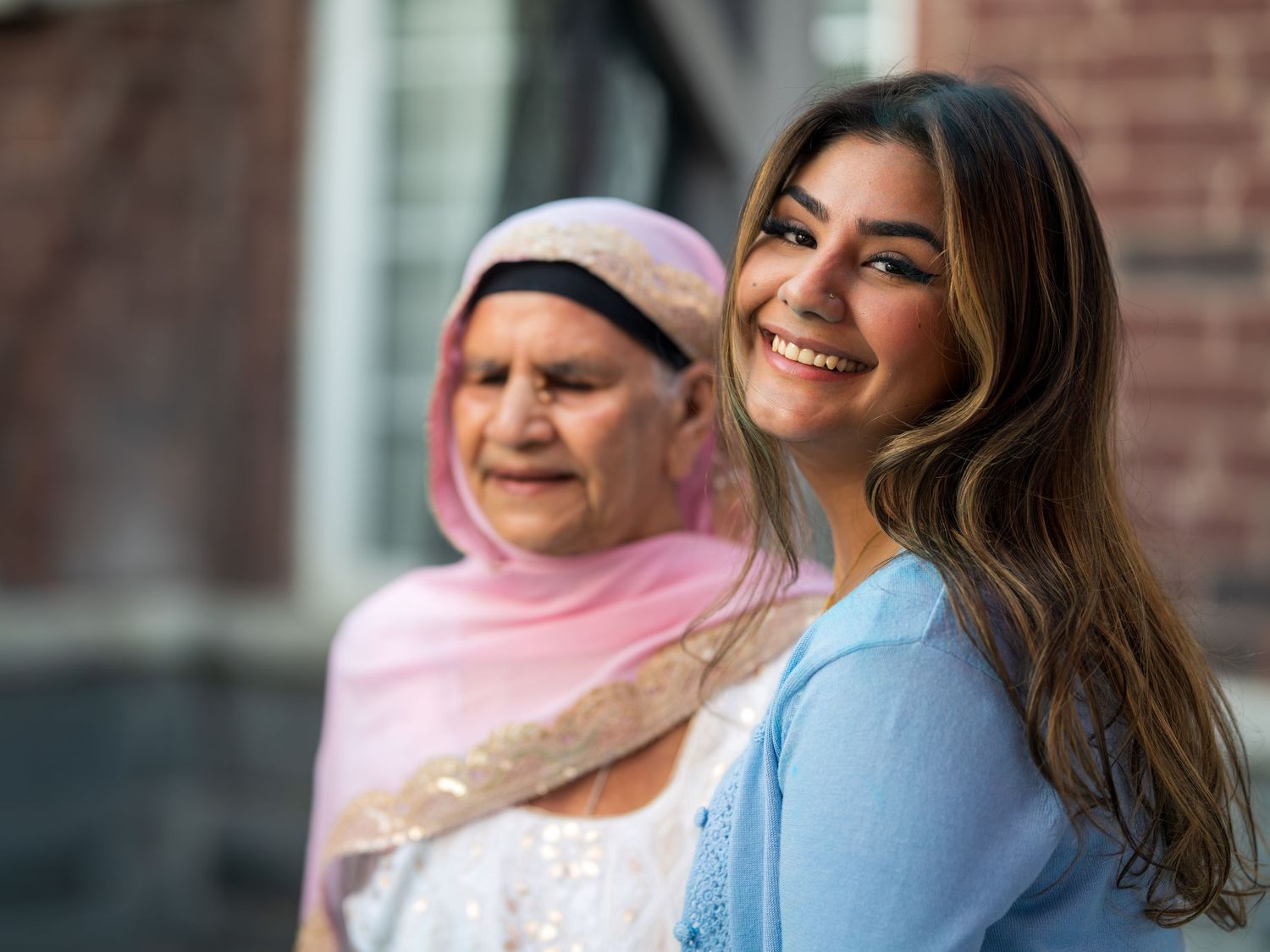 A smiling young woman in blue stands in front of an older woman wearing a pink hijab outdoors.