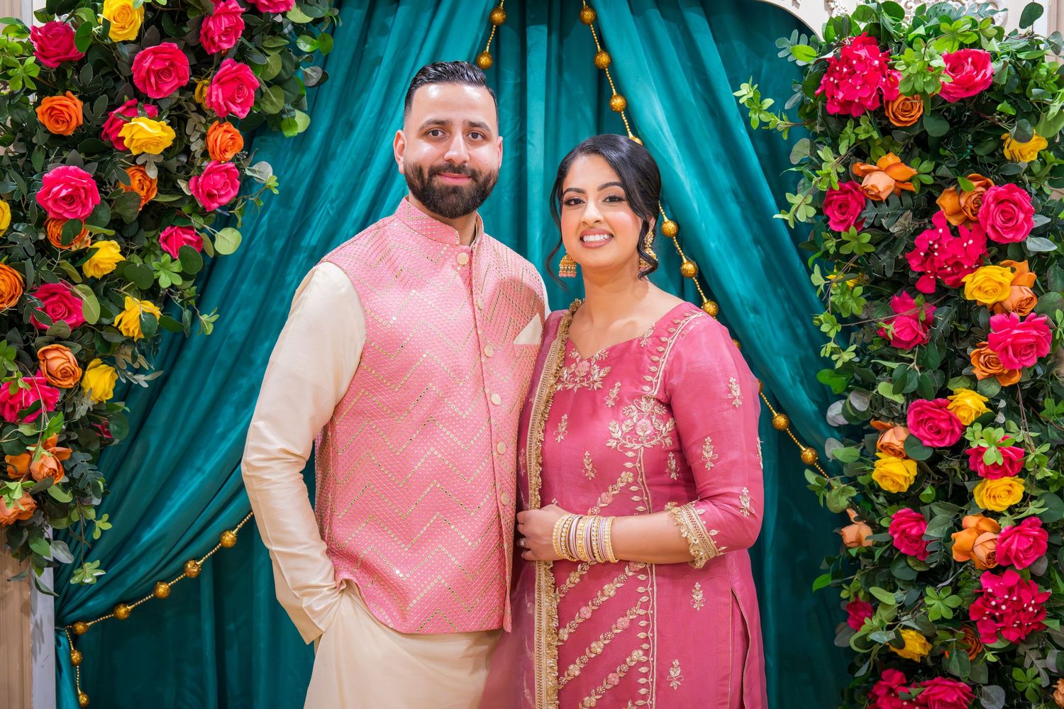 South Asian couple in coordinating pink traditional attire posing at colorful floral arch backdrop.
