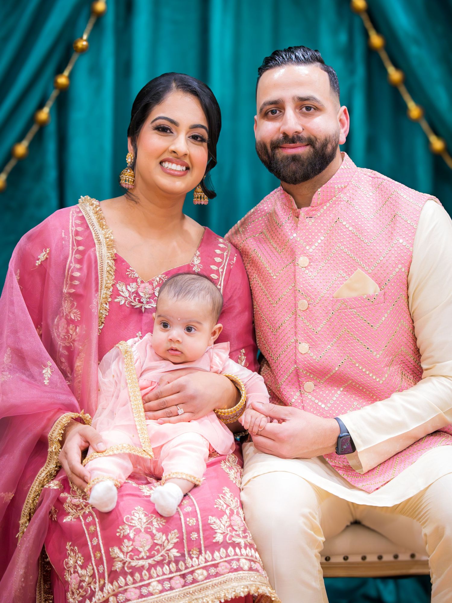 Smiling South Asian couple in pink traditional Indian outfits holding baby at decorated backdrop.