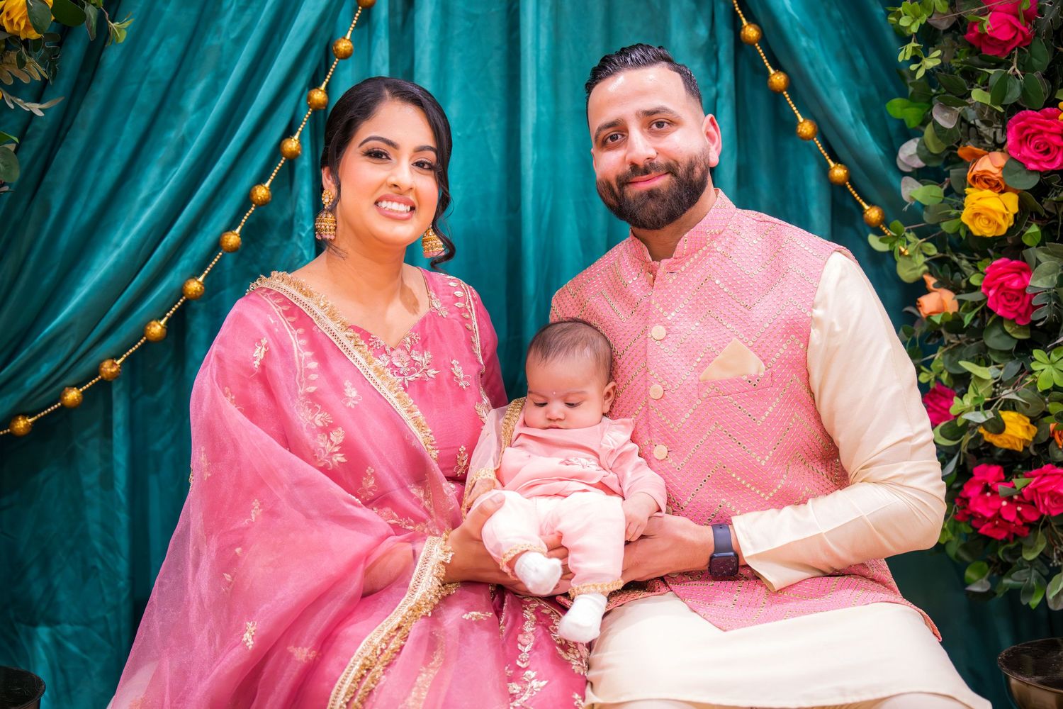 Smiling Indian couple in pink traditional attire holding newborn baby at colorful floral decorated celebration backdrop.