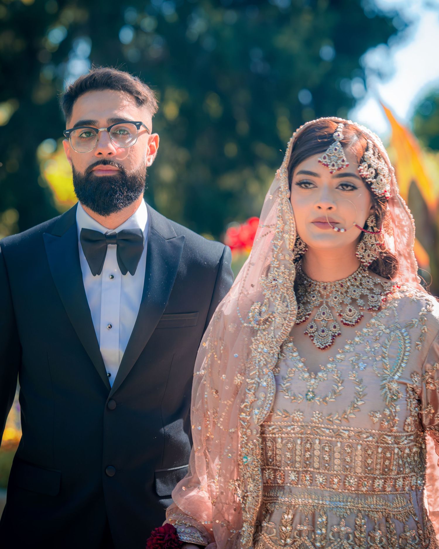 A wedding couple in traditional South Asian attire pose together against a sunlit garden backdrop.