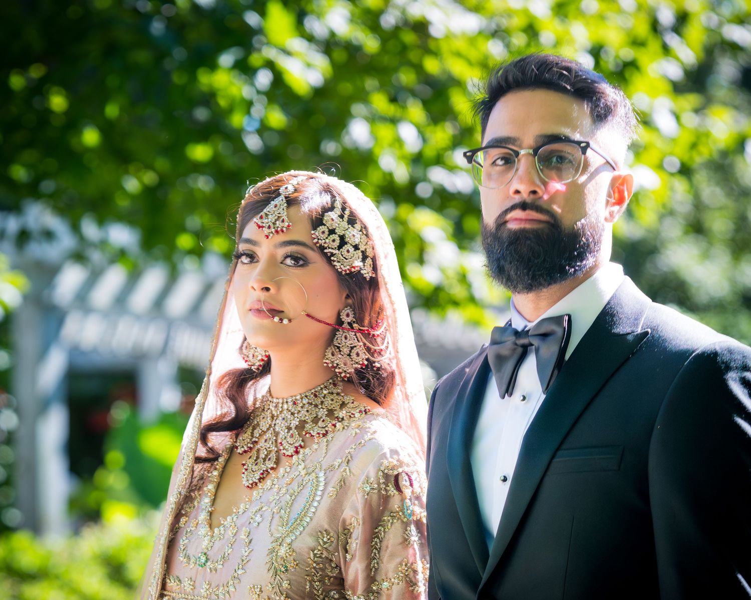 Elegant South Asian wedding portrait featuring traditional bridal dress and groom in tuxedo with garden backdrop.