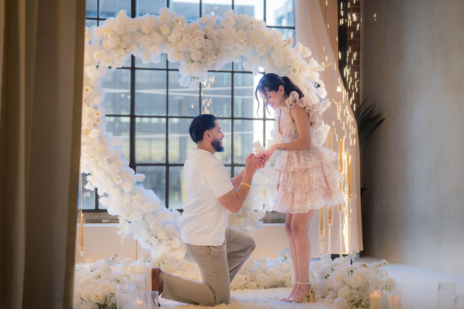 Man proposes on one knee inside white floral heart arch with fairy lights.