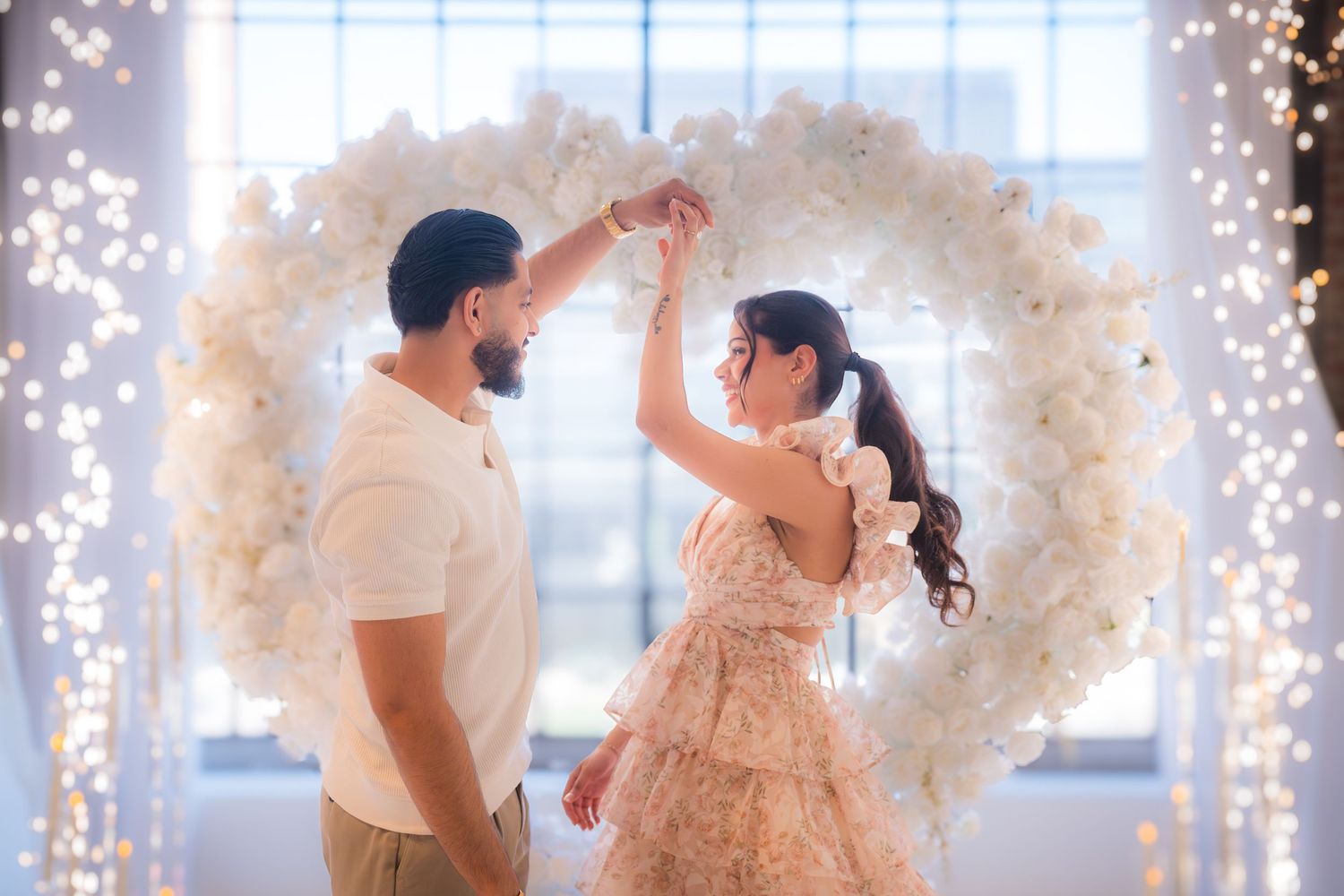 Couple dancing together under fairy lights in front of large white floral heart arch decoration.