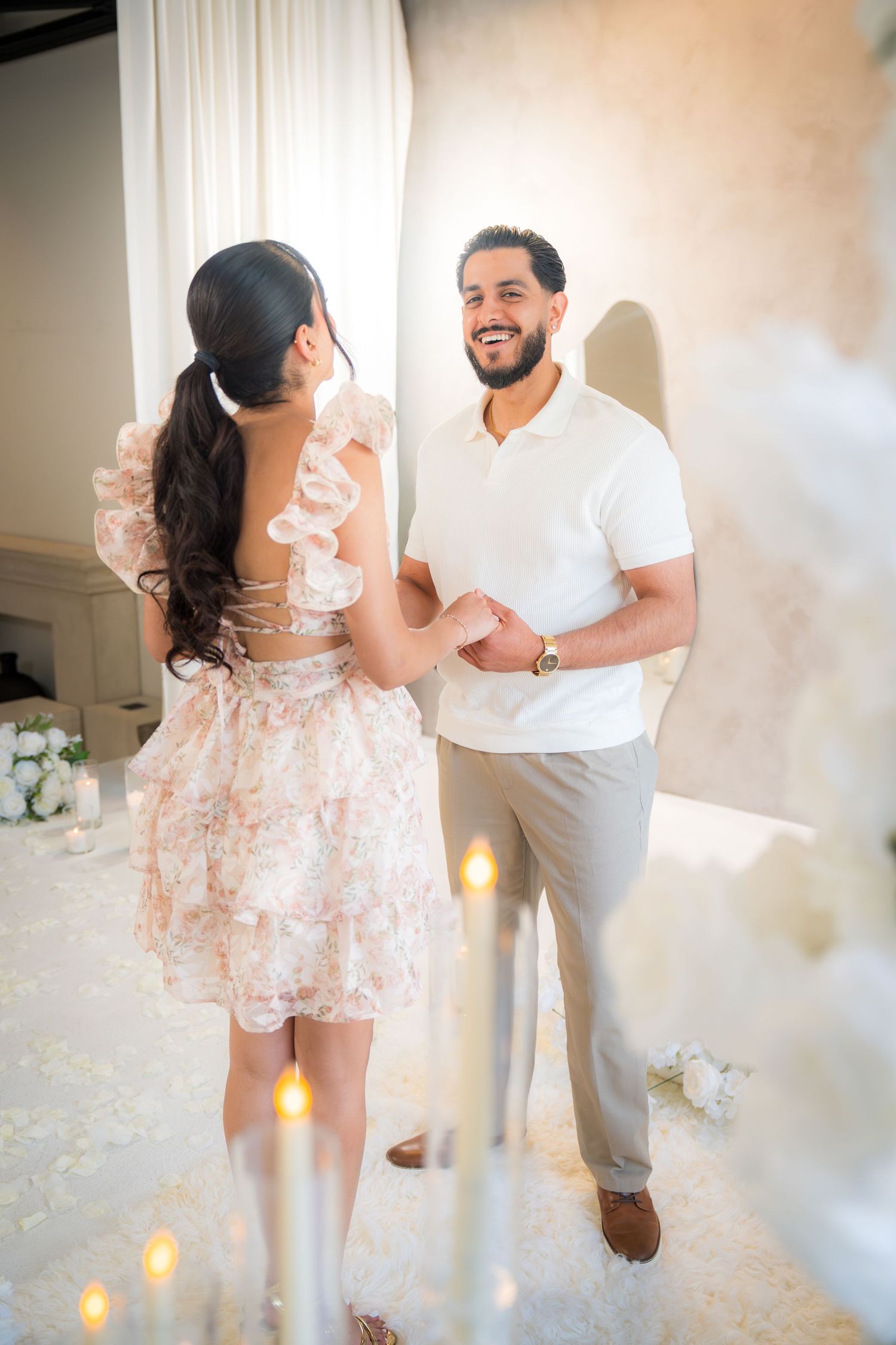 Smiling couple holding hands in an elegant candlelit white room during a romantic proposal setup.