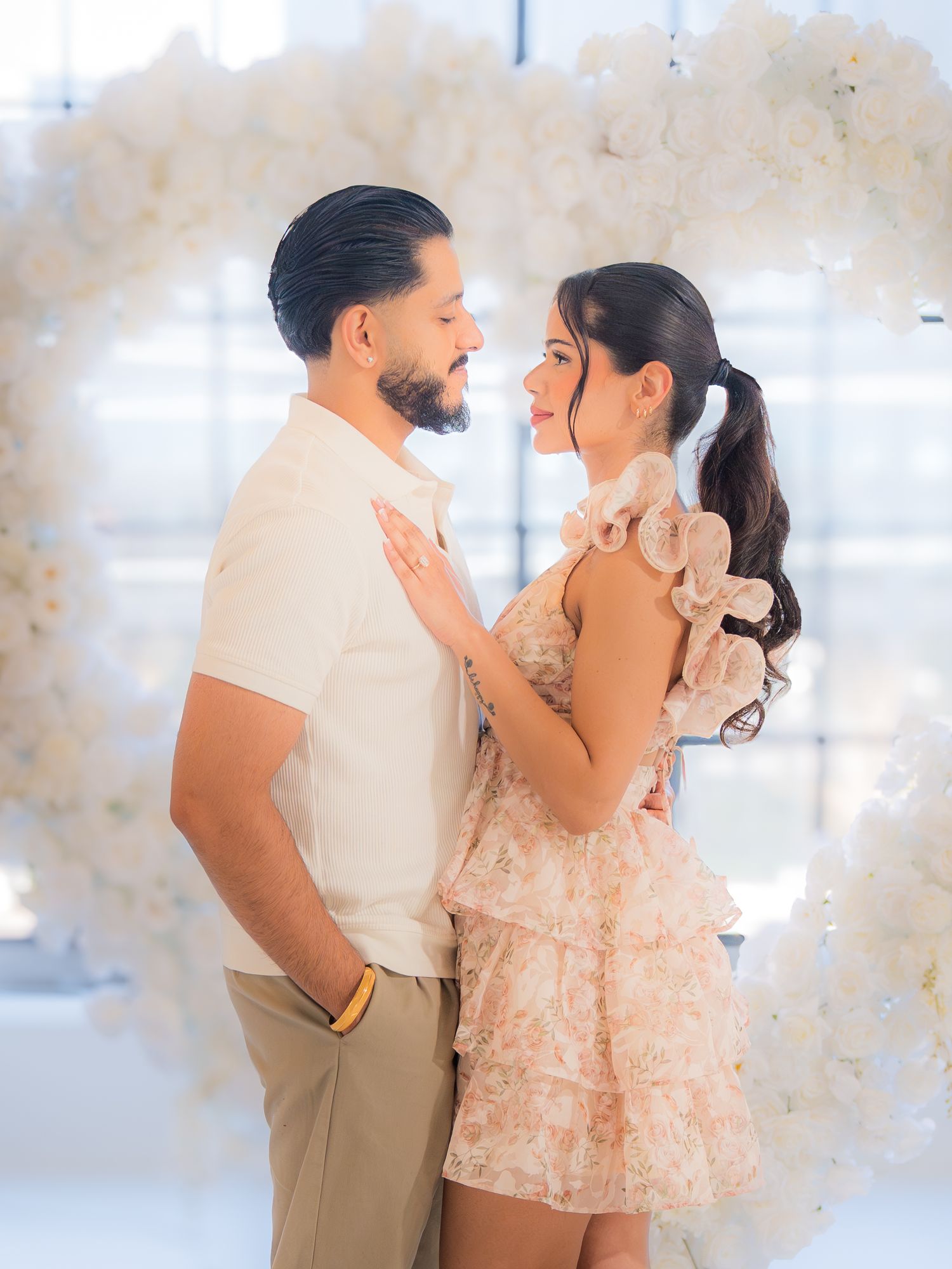 Couple gazing lovingly at each other in front of a large white floral heart arch backdrop.