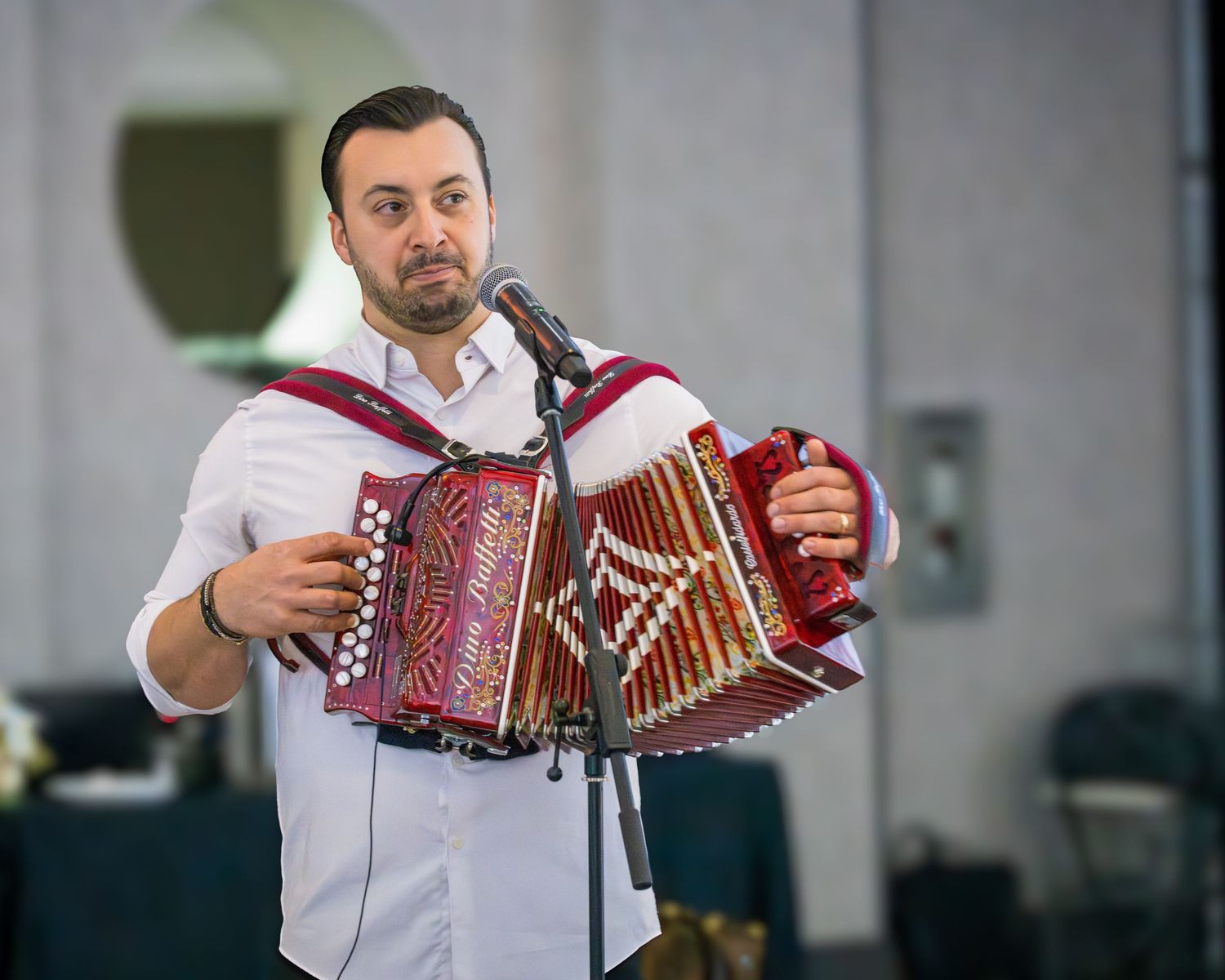 Male accordion player performing at a formal event, playing a red button accordion at a microphone stand.