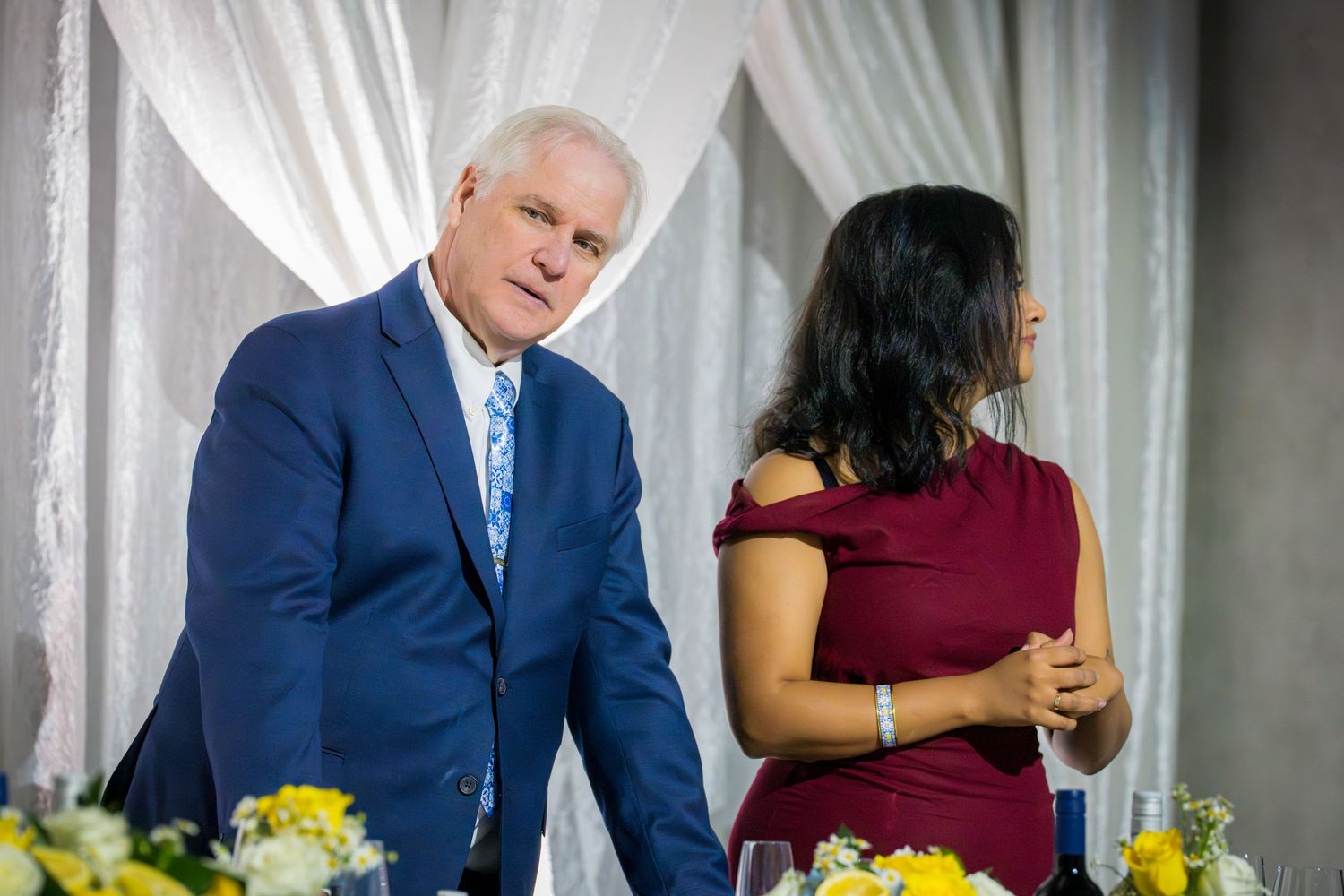 Man in blue suit standing beside woman in red dress near yellow and white floral centerpieces at anniversary event.