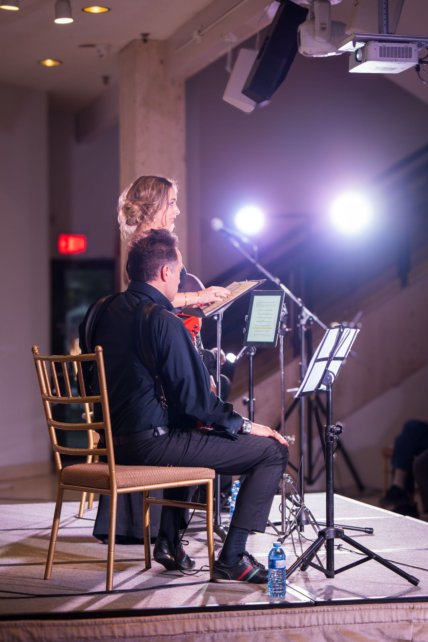 Two musicians perform on stage, a woman playing violin and a man seated, illuminated by bright stage lights.