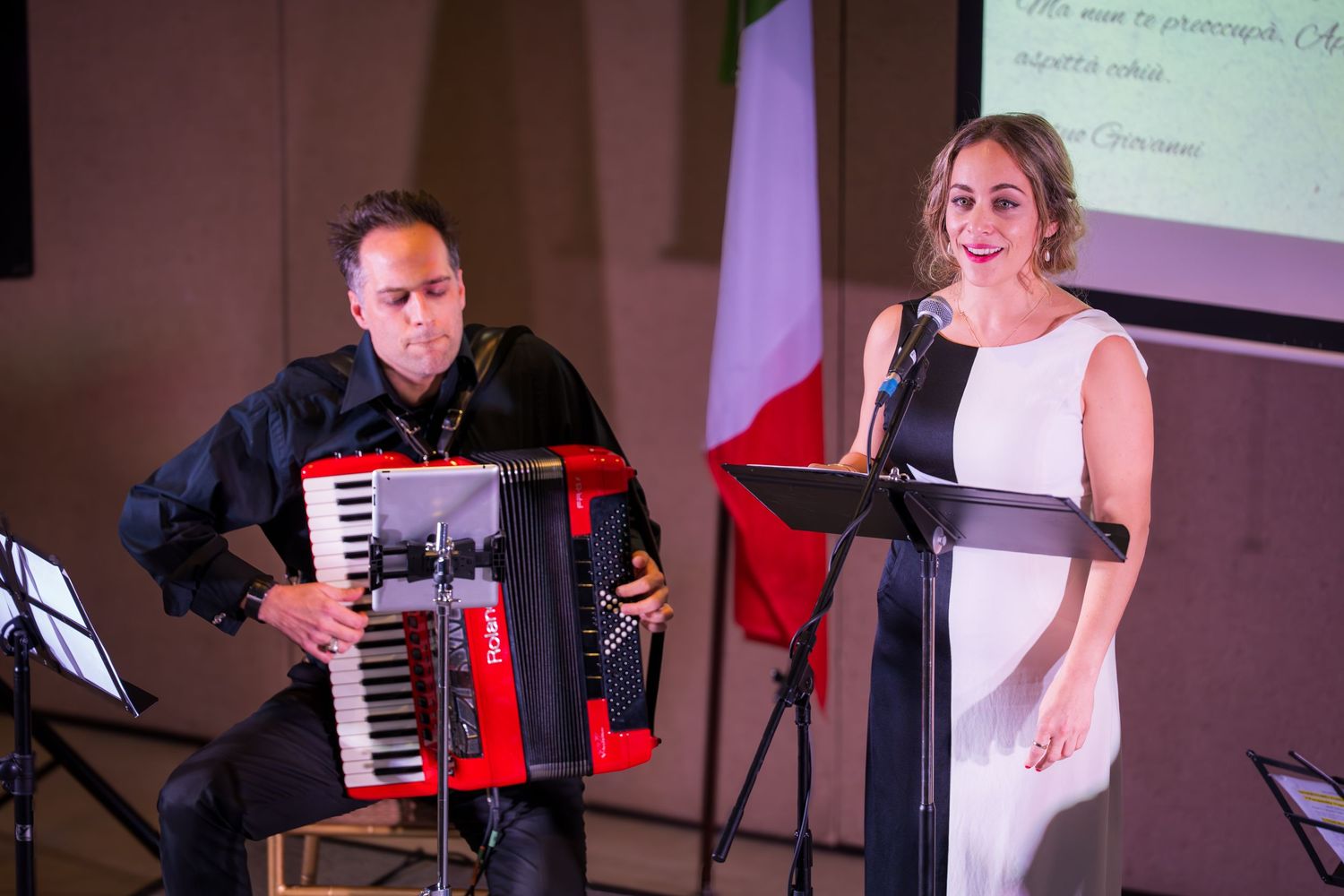 Male accordionist playing a red Roland accordion beside a female singer at a microphone stand on stage.
