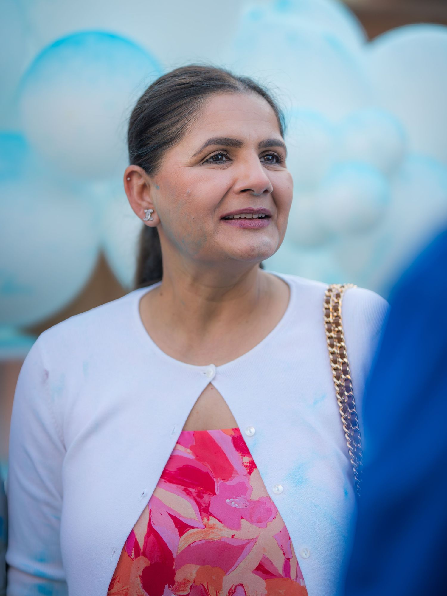 A woman in a white cardigan and floral dress smiling near blue and white balloons at an outdoor event.