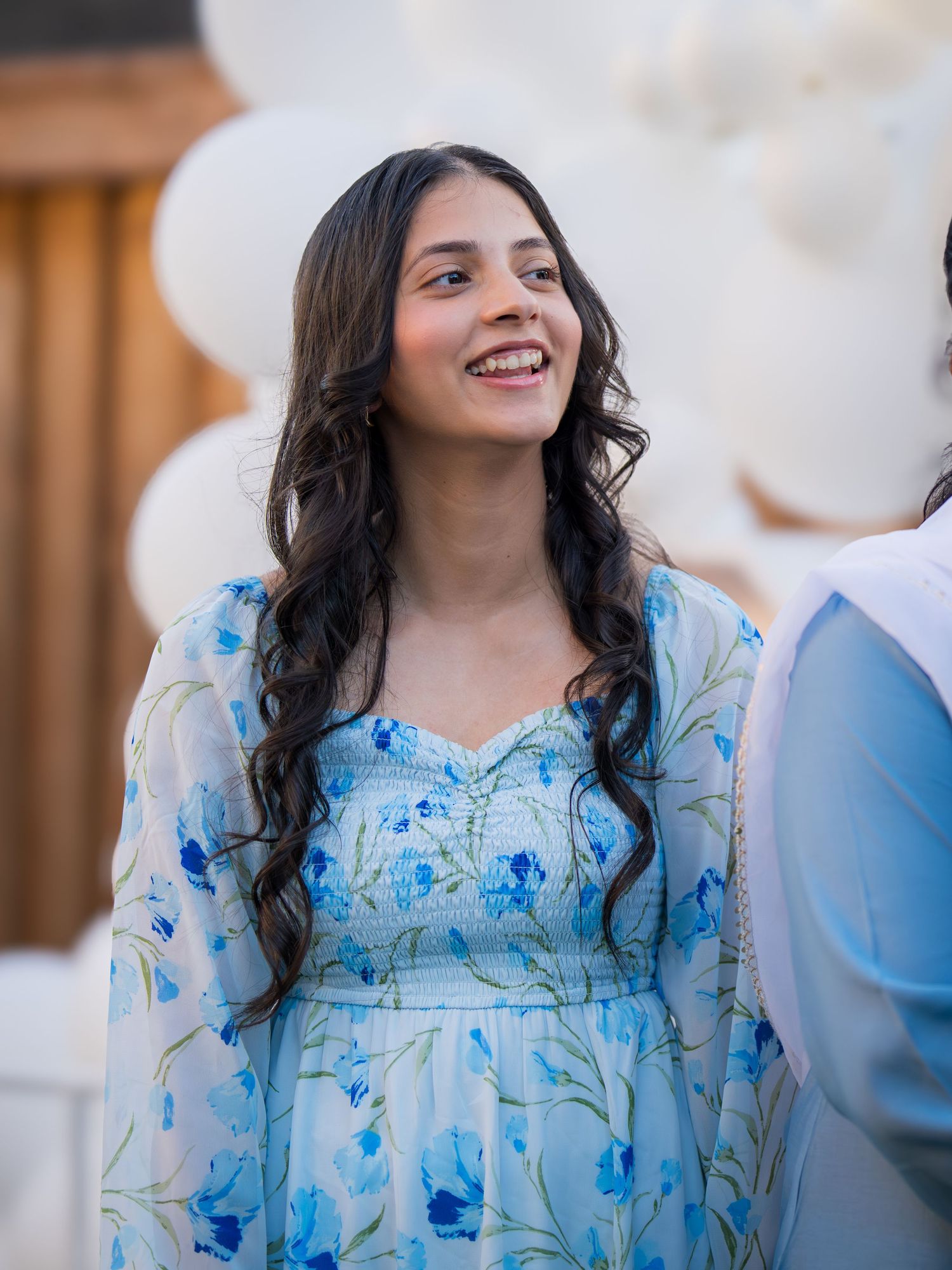 Young woman with long dark hair smiles in blue floral dress at outdoor celebration with white balloons.