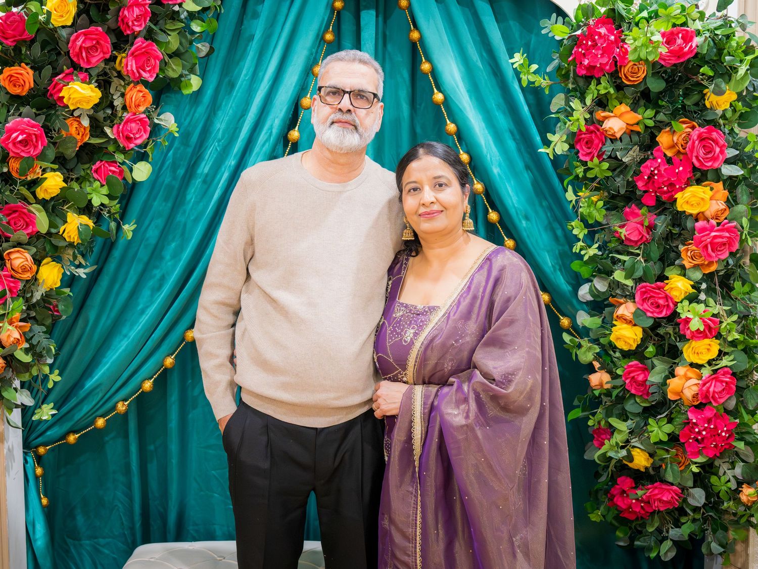 Elegant older couple smiling together at a vibrant floral South Asian celebration backdrop.