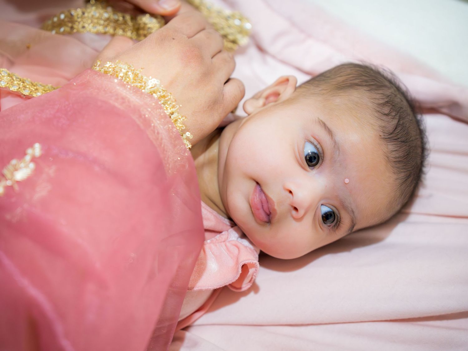 Adorable baby girl in pink traditional Indian outfit with gold jewelry and bindi, lying on a pink surface.