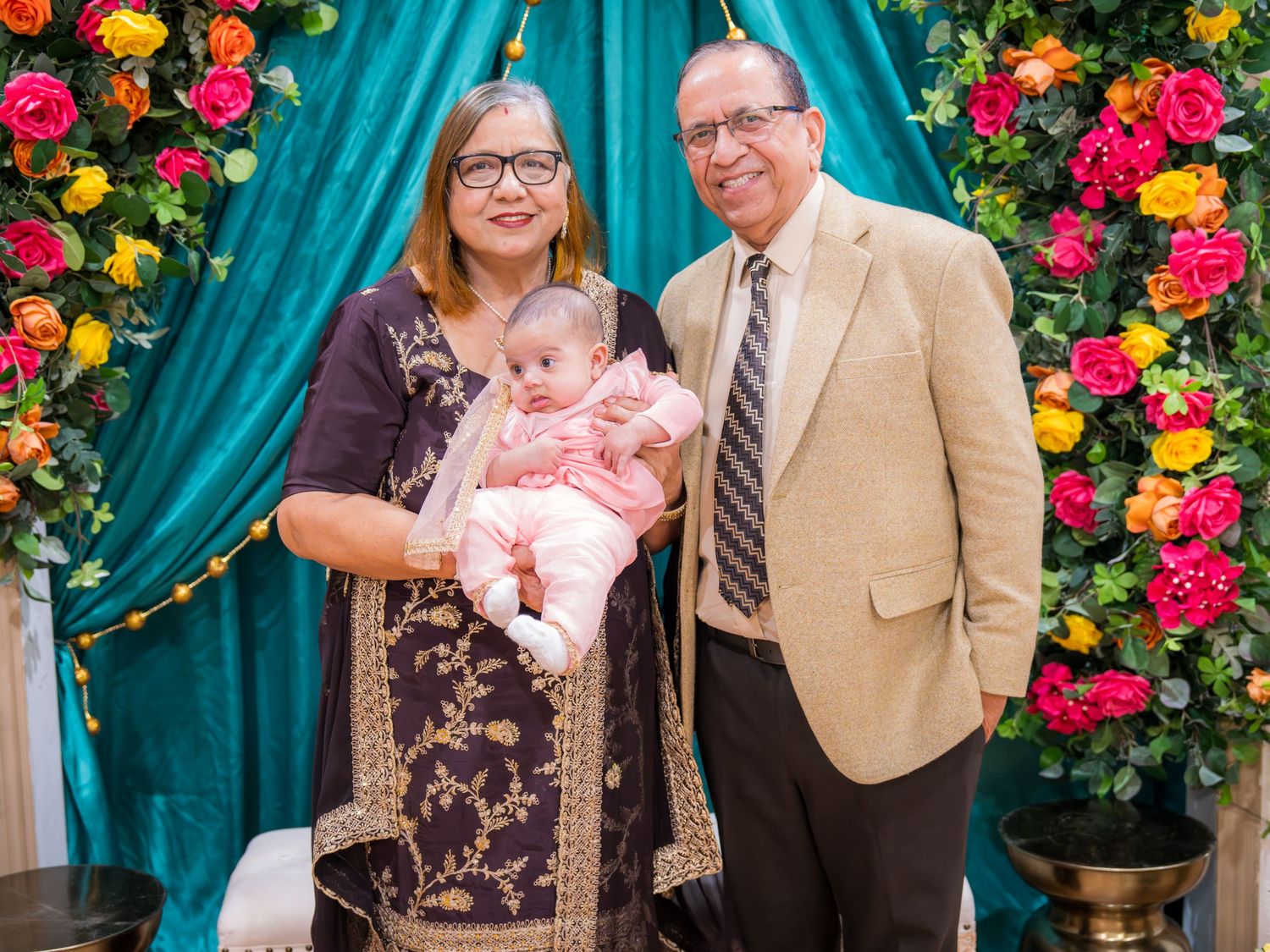 An older couple in formal wear holds a newborn baby in front of a vibrant floral and teal backdrop.