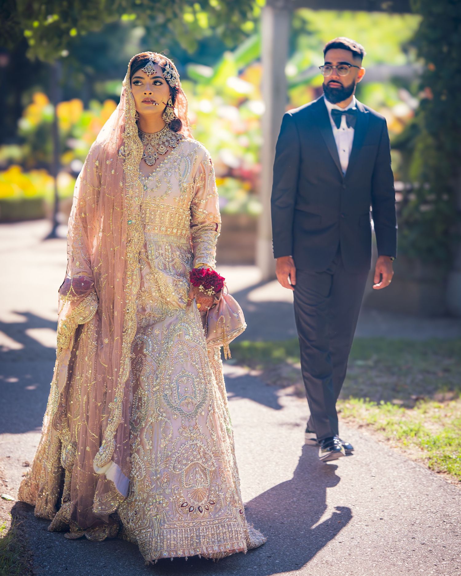 Couple strolling on a sunlit path, bride in ornate peach dress and groom in black suit.