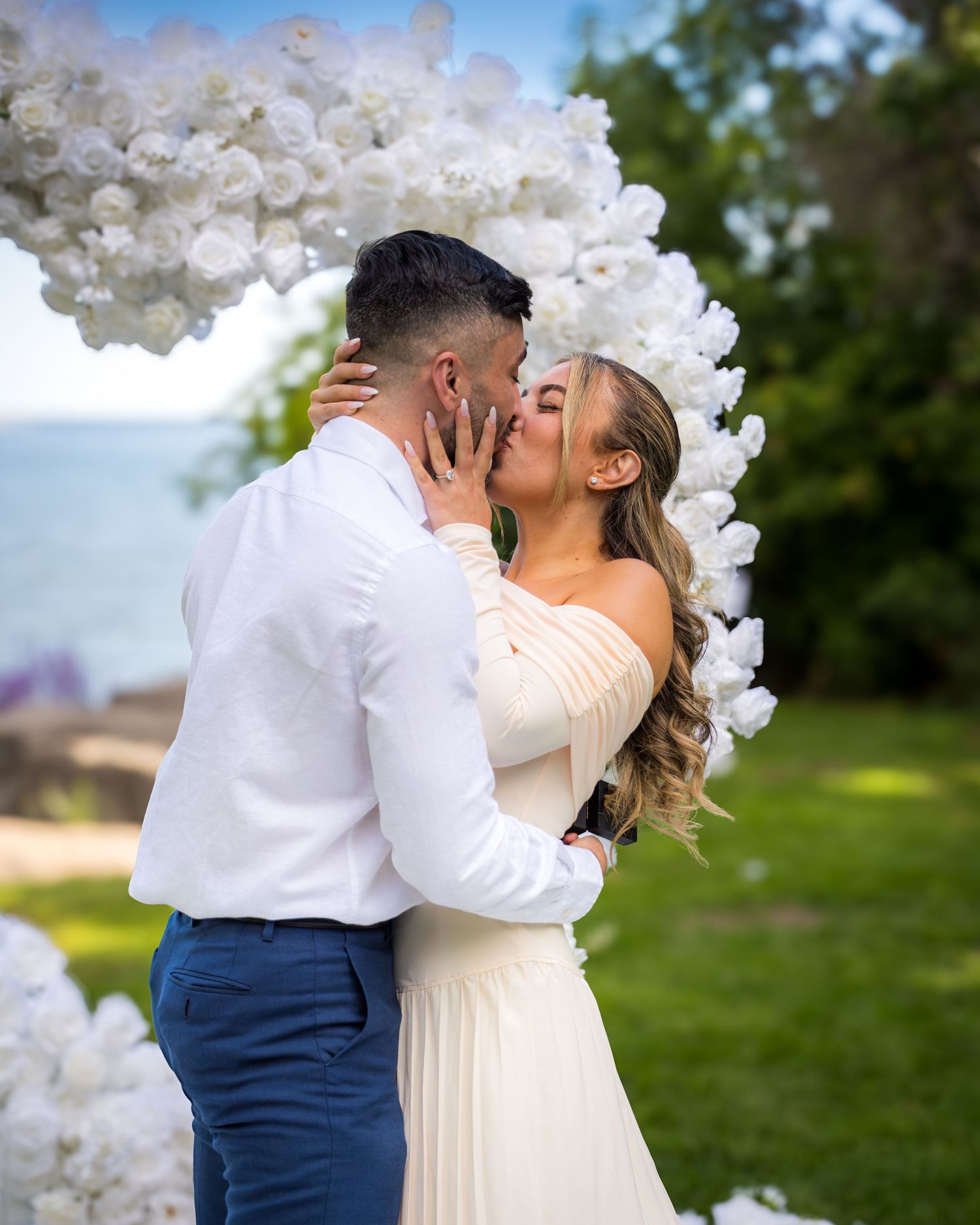 Couple sharing first kiss under white floral arch during outdoor ceremony.