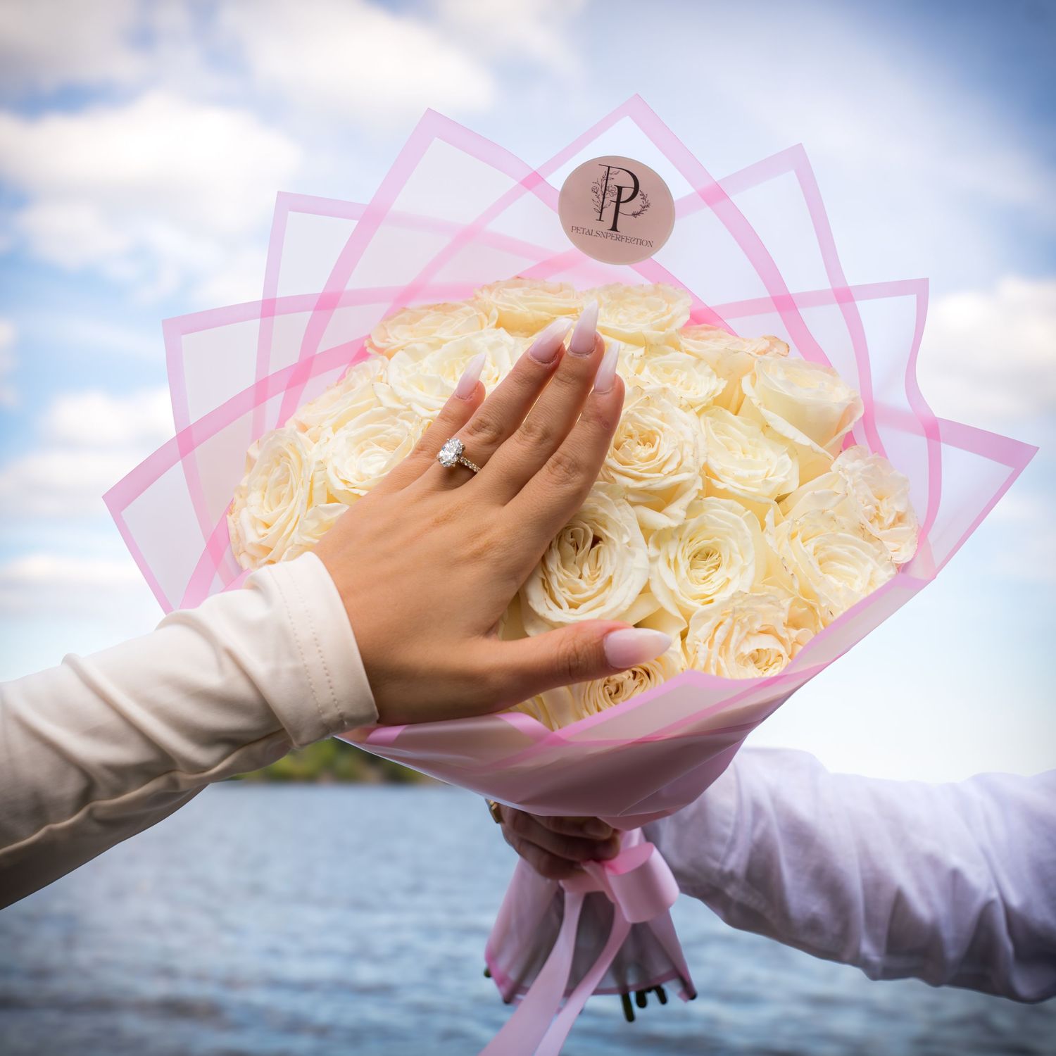 Close-up shots of hands holding a pink-wrapped bouquet of white roses against an ocean backdrop.