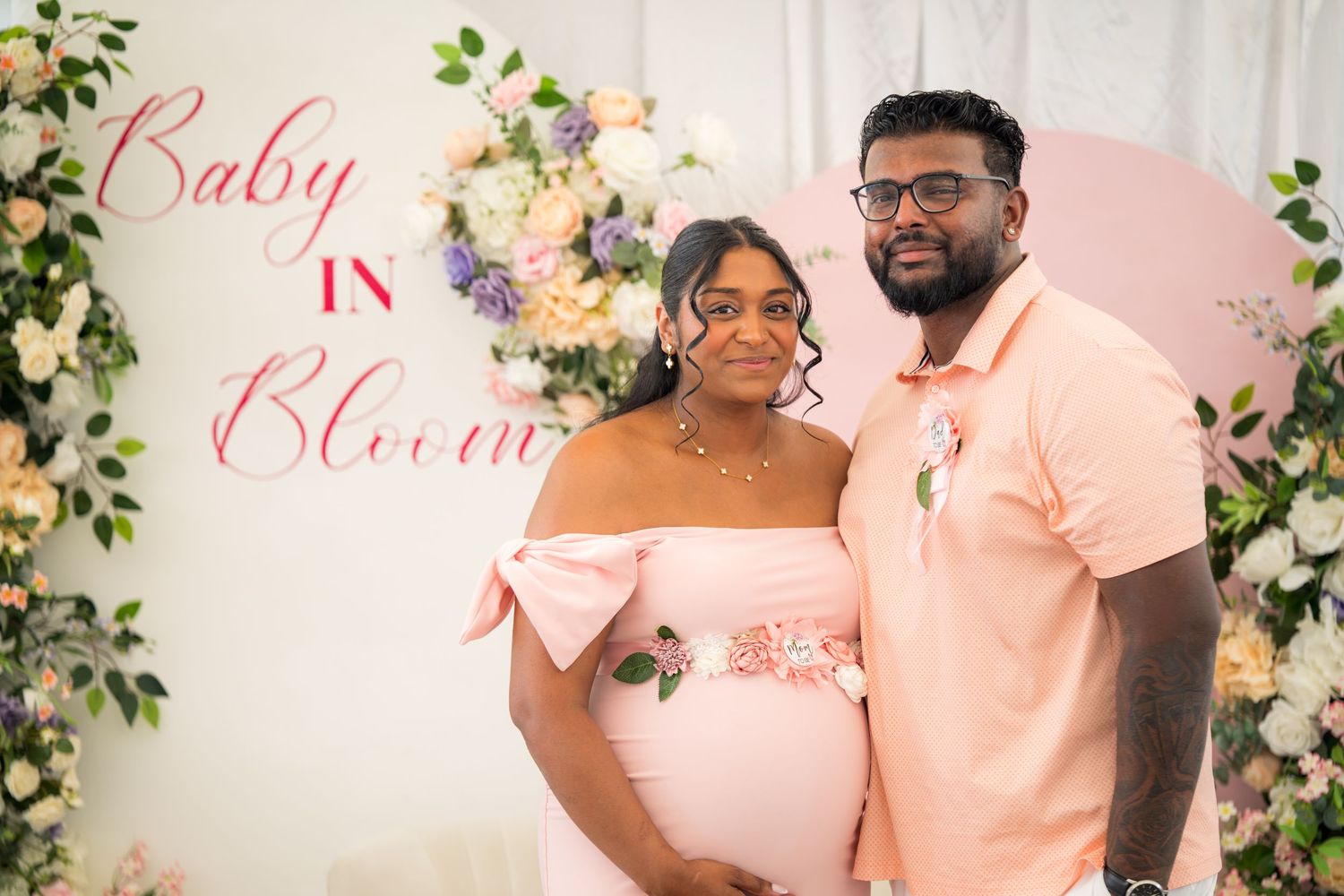 Couple celebrates at baby shower with pink and white floral backdrop and coordinated outfits.