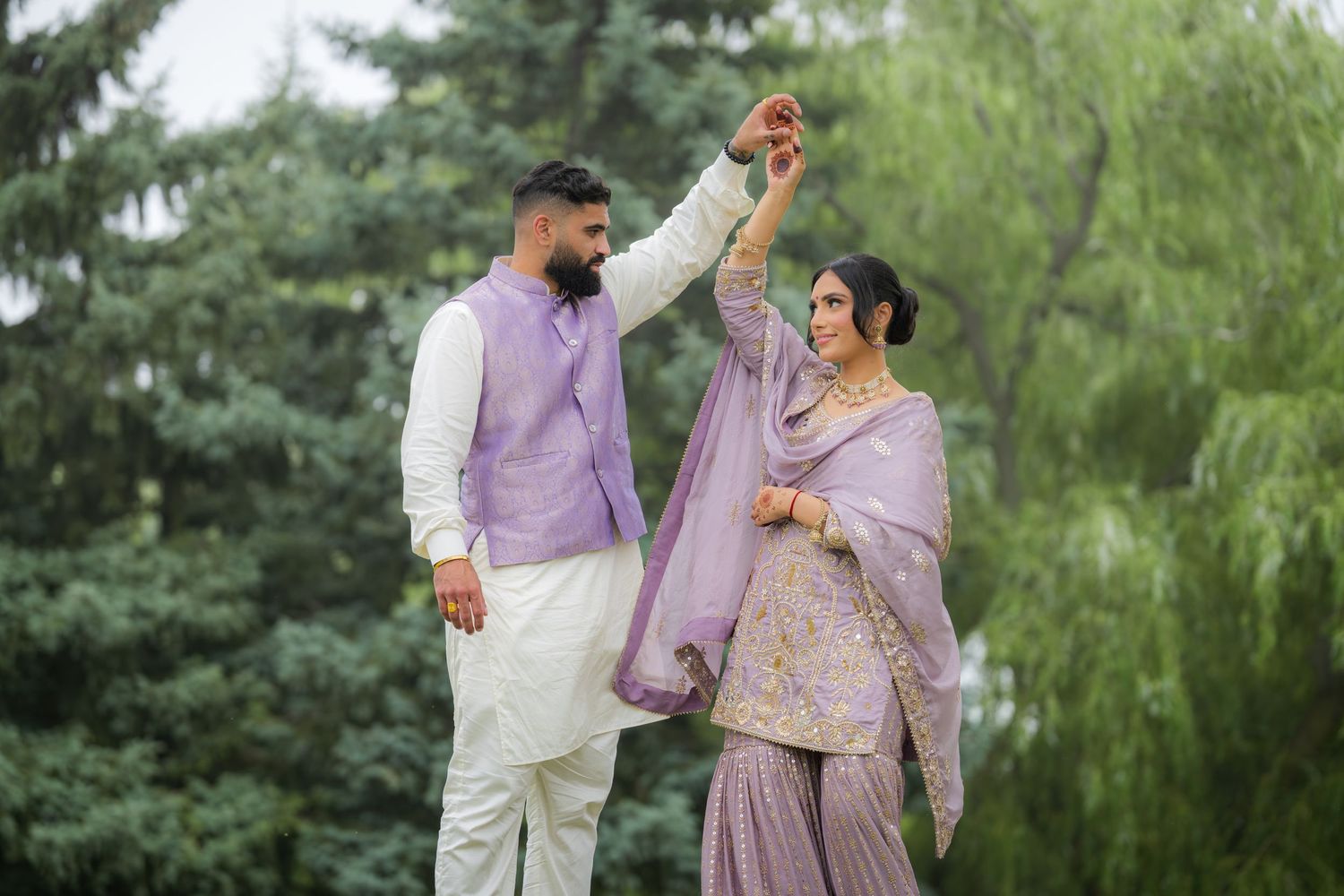 A couple in traditional South Asian attire dance together against a backdrop of evergreen trees.