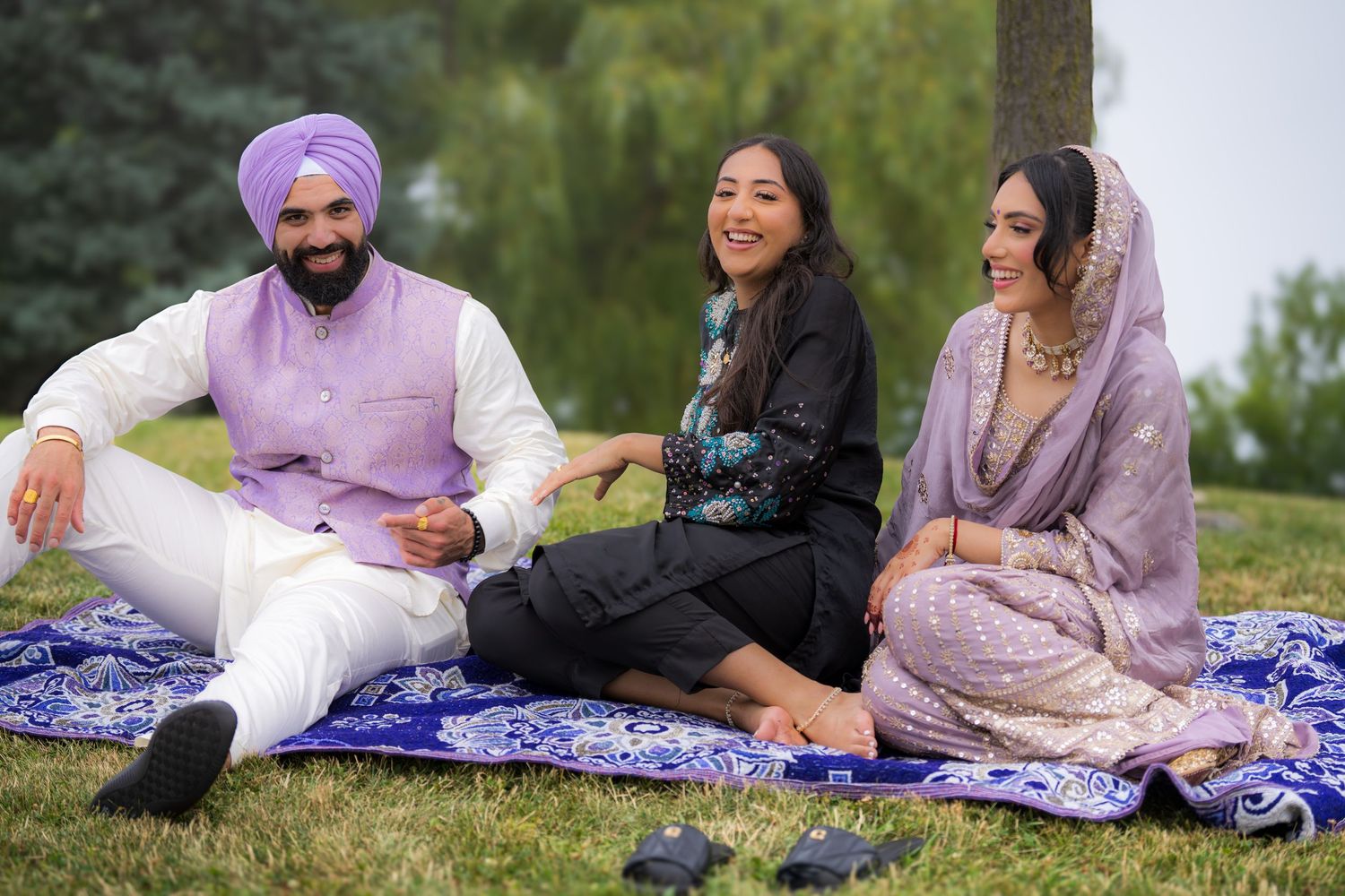 Three people lounge casually on a blue patterned blanket outdoors wearing traditional Indian-inspired attire.