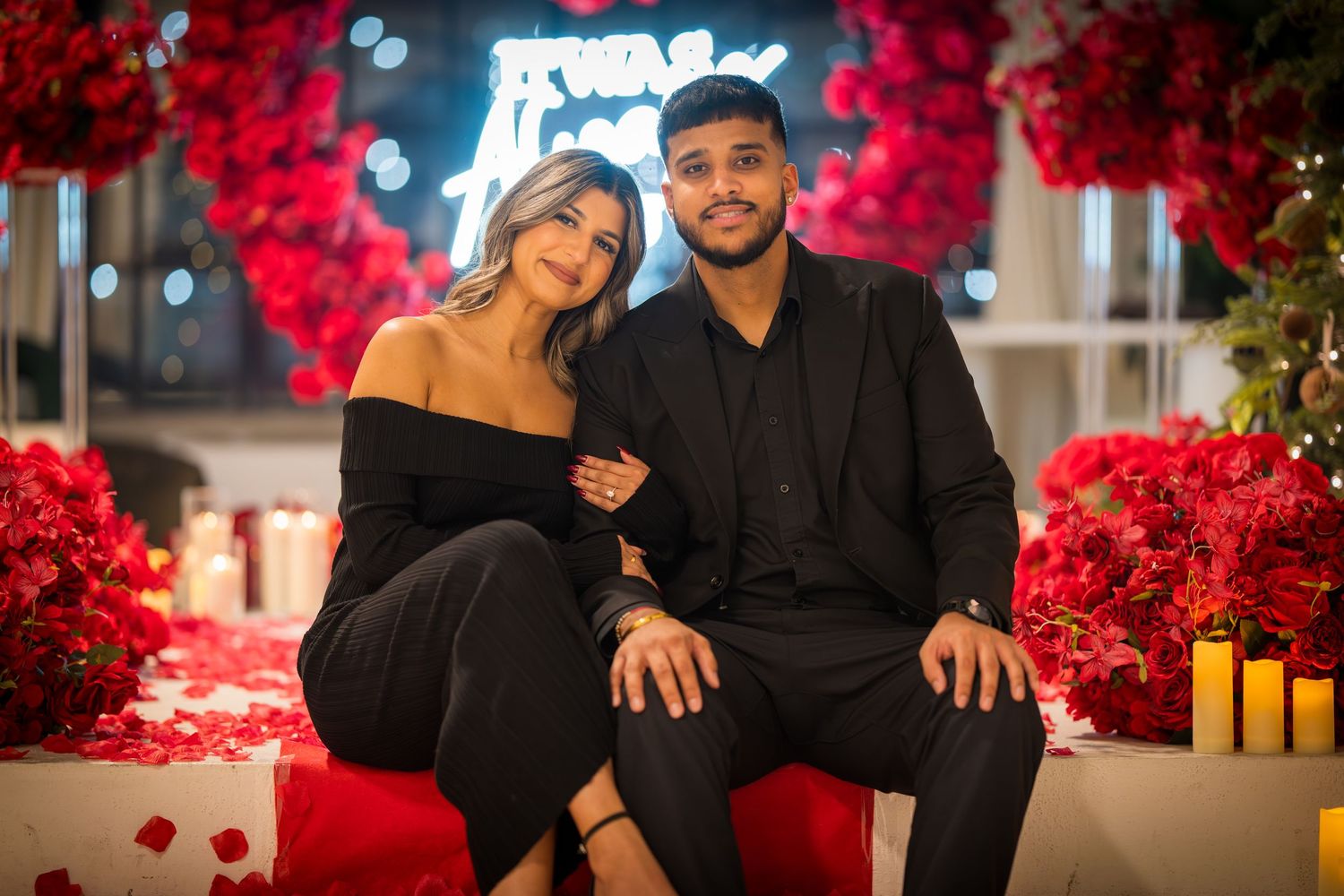 Couple sits amid romantic setting with red roses, candles, and soft lighting.